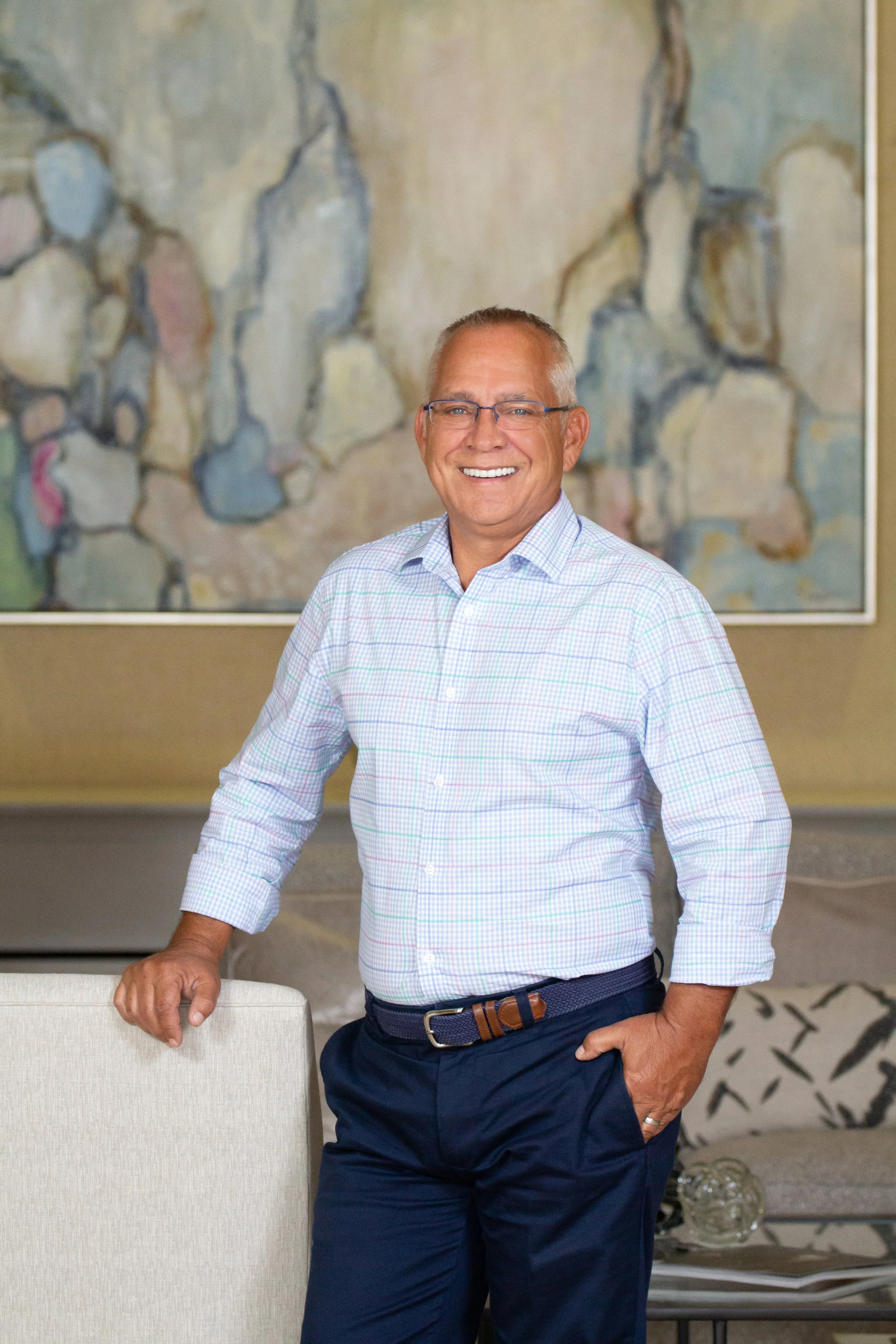 A smiling middle-aged man with glasses, wearing a blue checkered shirt and navy pants, standing in a room with abstract wall art in the background.