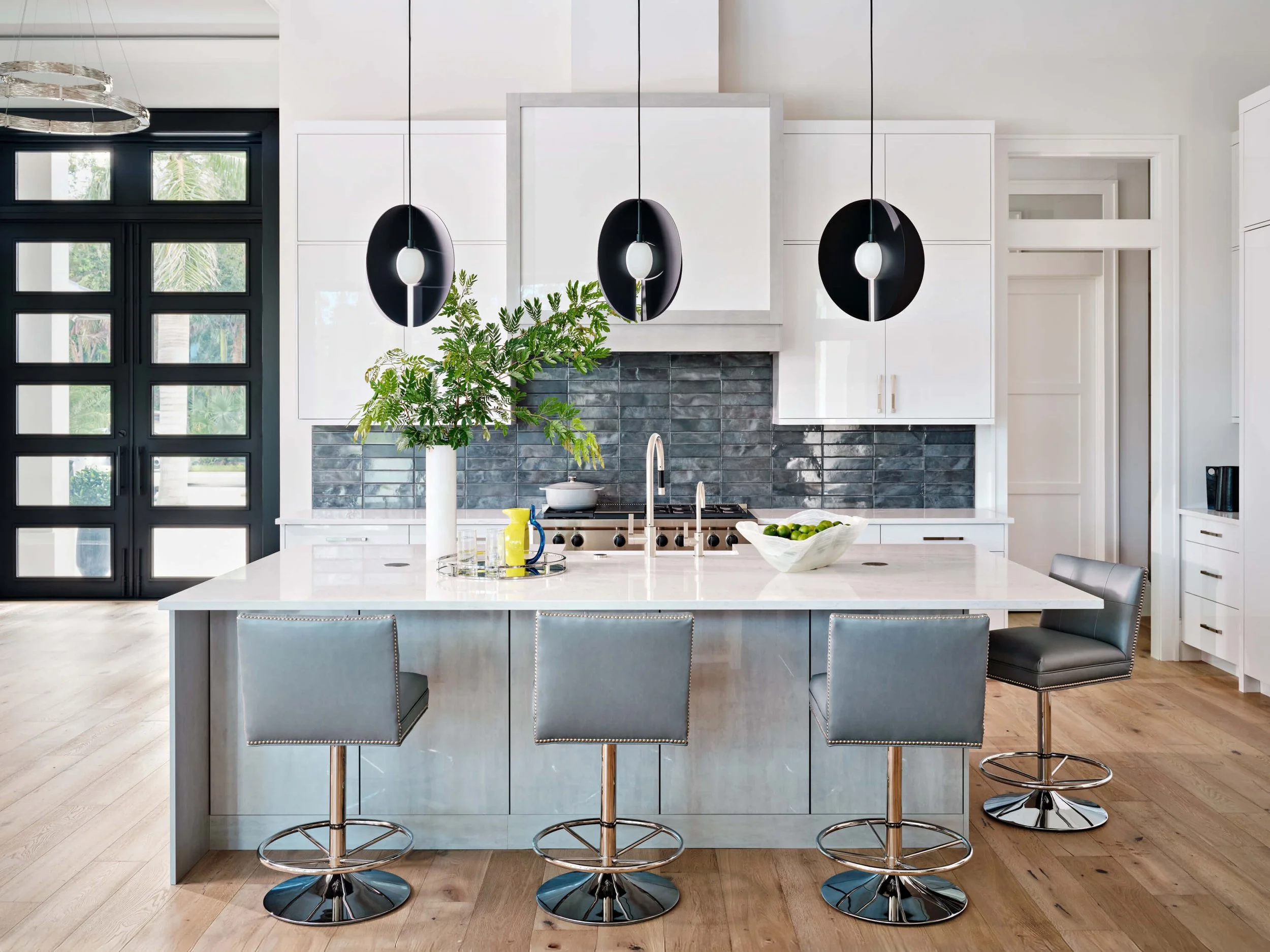 Modern kitchen with white cabinets, black backsplash, a large white island with bar stools, pendant lights, and a potted plant near the window.