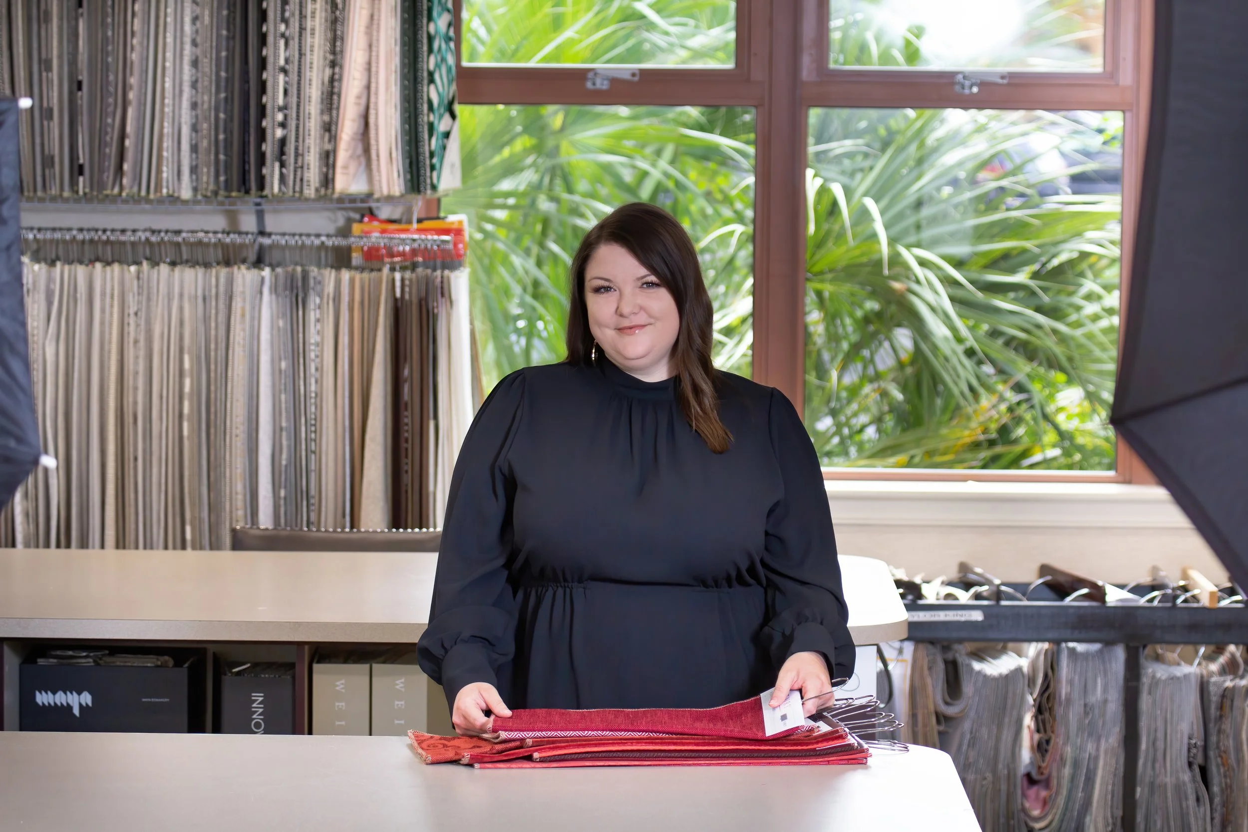 A woman in a black dress standing behind a counter, holding fabric samples in fabric store with shelves of fabric rolls and sample books, large window with greenery outside.