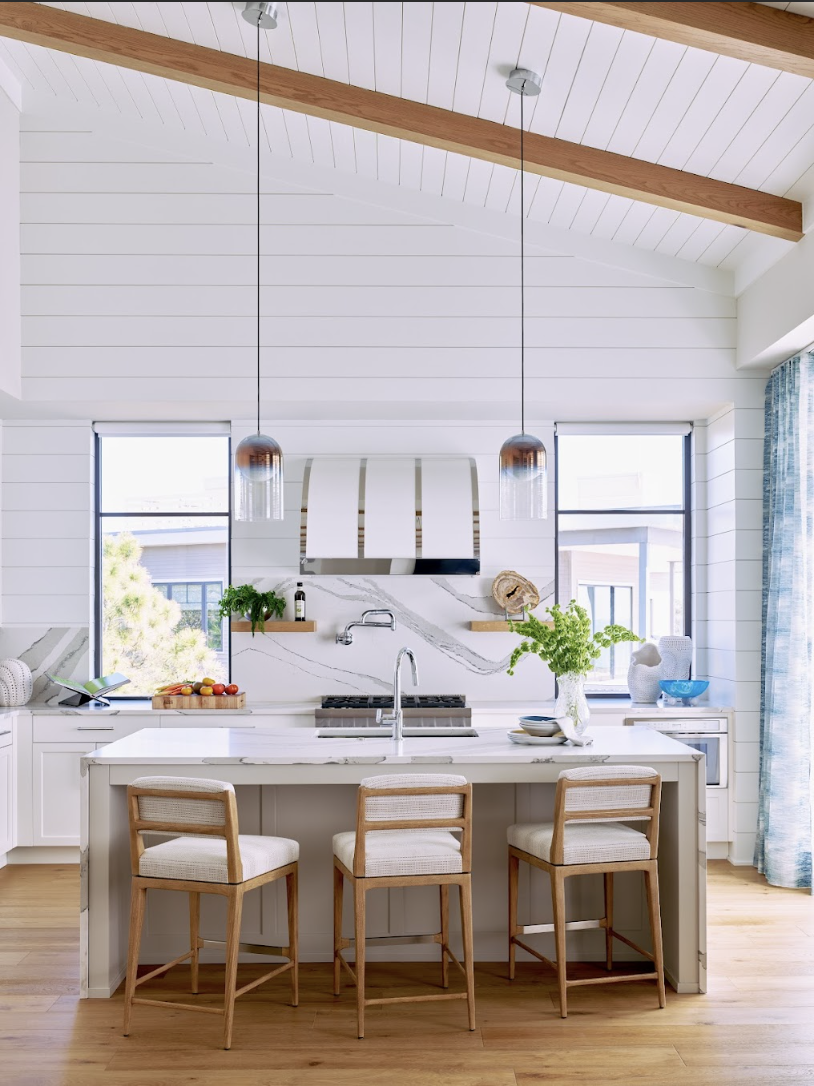 Bright, modern kitchen with white cabinetry and marble backsplash, three wooden barstools at a kitchen island, two pendant lights, and large windows letting in natural light, with wooden beams on the ceiling.