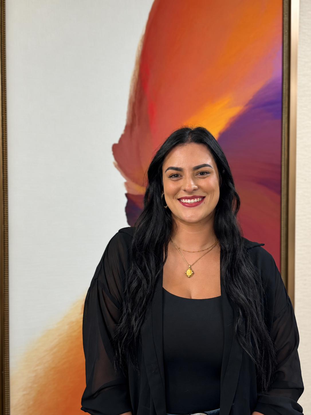 A woman with long dark hair, wearing a green cardigan over a black top and jeans, smiling while standing in front of an abstract painting with blue, green, and beige tones, and a decorated background with vases and plants.