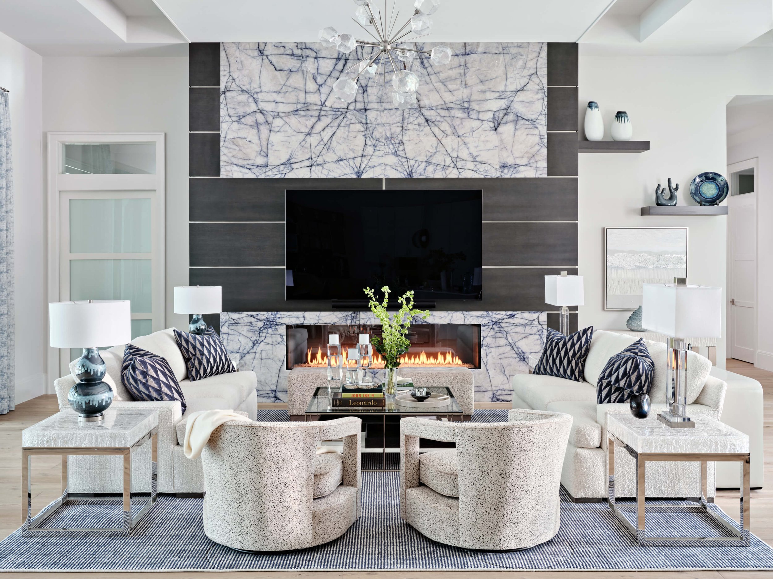 A modern living room scene with white and navy decor. It features a wall-mounted flat-screen TV, a fireplace below, and a marble accent wall above. The room has white sofas with navy patterned pillows, glass and metallic side tables, a glass coffee t