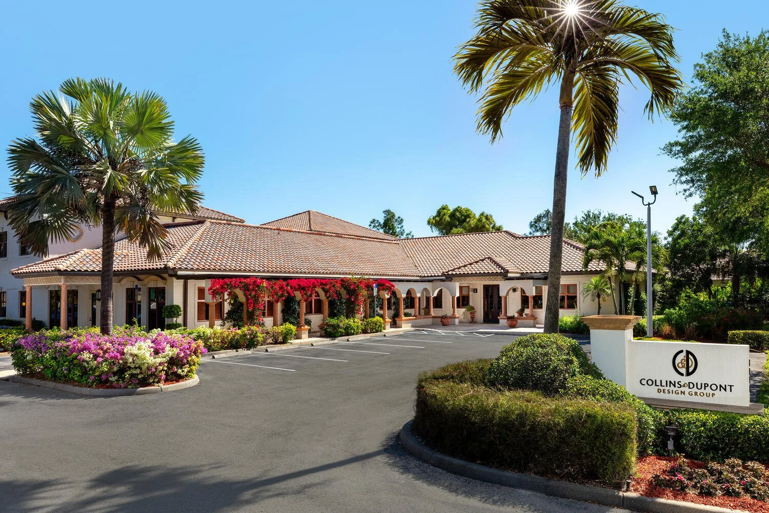 A Mediterranean-style building with a red-tile roof and arched windows, surrounded by lush greenery and colorful flowers, with a parking lot in the foreground and a sign that reads 'Collins Dupont Design Group'.