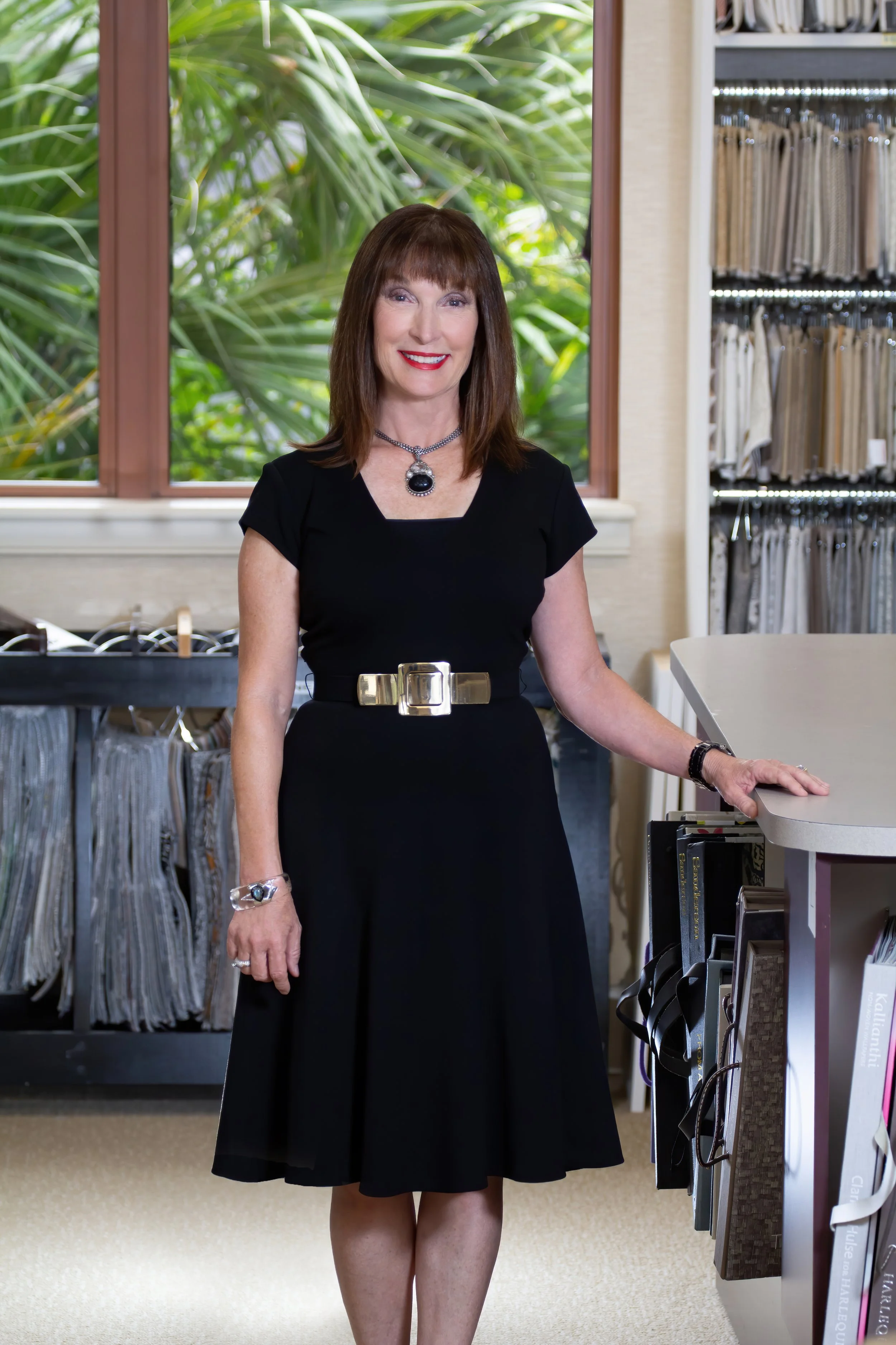 A woman with shoulder-length brown hair and bangs, wearing a black dress with a gold belt, standing inside a fabric store. She is smiling and wearing a large necklace with a black pendant, a bracelet, and rings. Behind her are fabric samples and a window with green foliage visible outside.