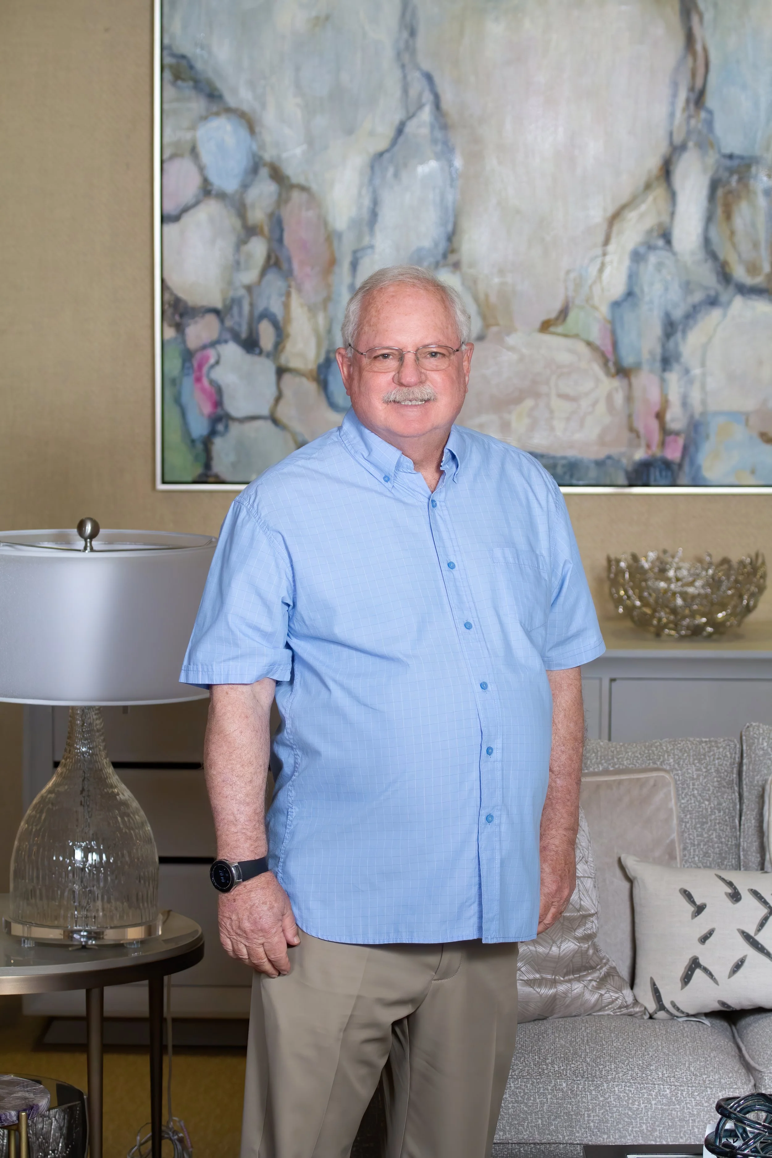 A man with gray hair, glasses, and a mustache wearing a light blue button-up shirt and beige pants standing in a living room with a colorful abstract painting behind him. The room features a table lamp, decorative bowls, and pillows on a sofa.