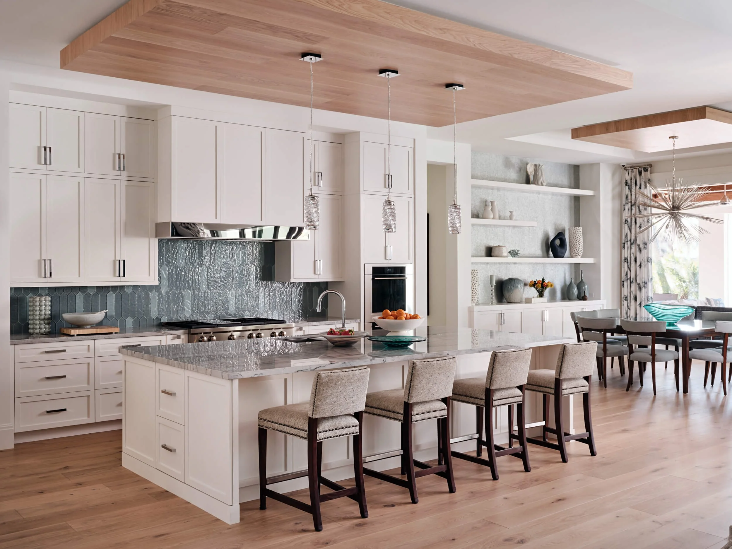 Modern kitchen with white cabinetry, a marble island, and a pendant light with glass details hanging above the island. There's a wooden ceiling feature, a textured glass backsplash, and open shelving with decorative items in the background.