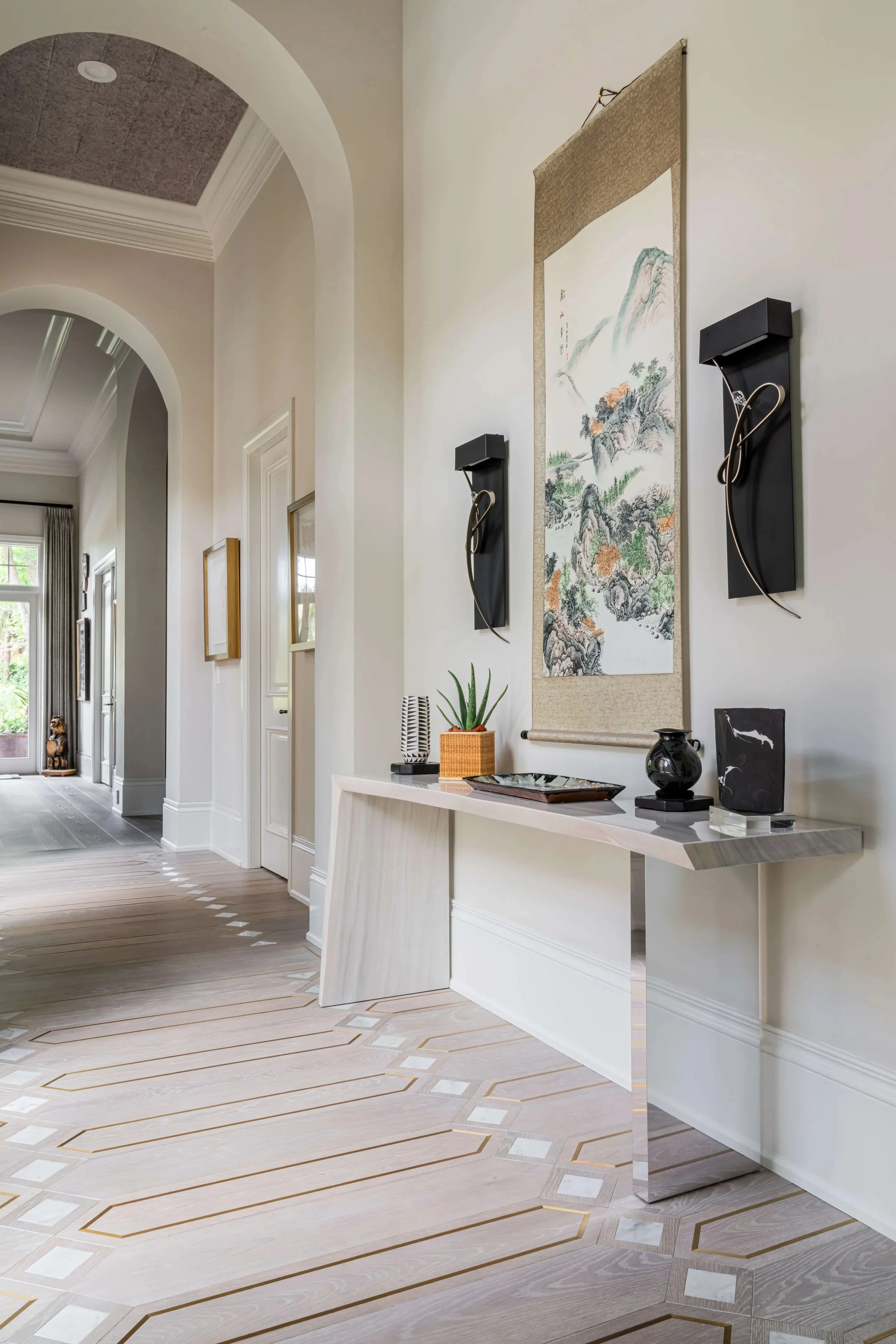 Interior hallway with light-colored wood flooring, decorative wall art, and modern wall sconces, with large arched doorways and windows bringing in natural light.