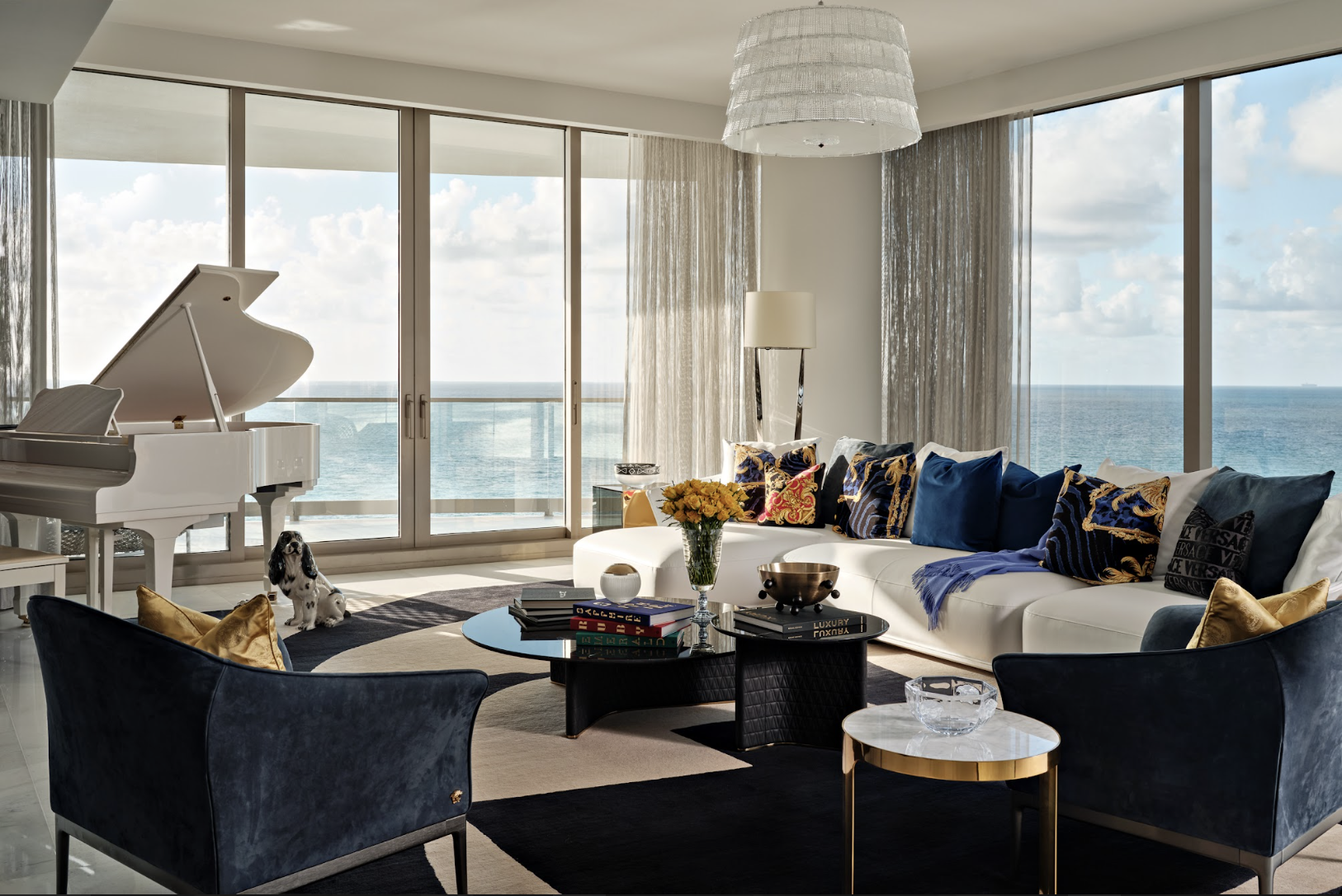 Living room with large floor-to-ceiling windows showing ocean view, white sofa with decorative pillows, black velvet chairs, a round coffee table with books, a flower vase, and a dog sitting near a white grand piano.