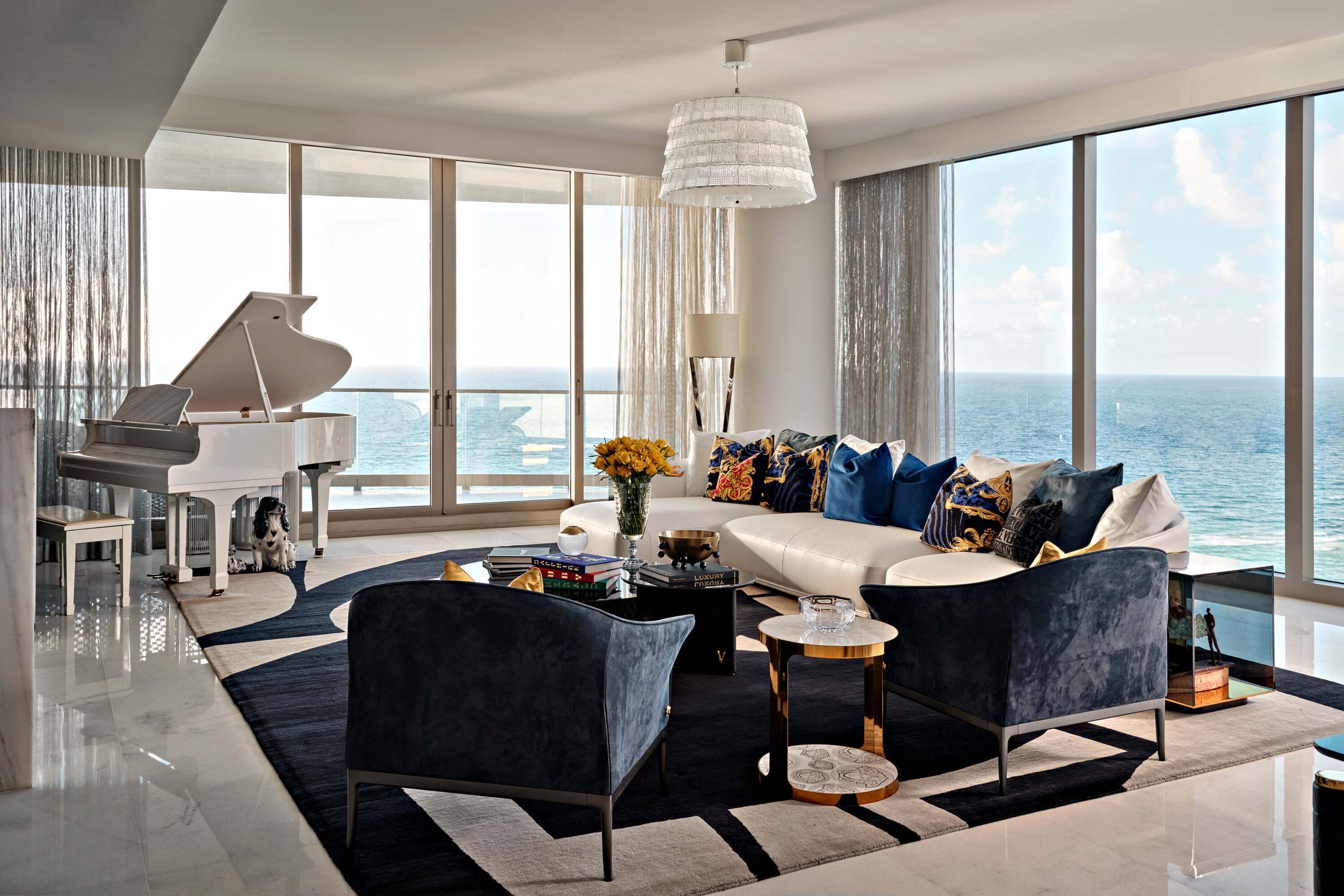 Living room with large windows overlooking the ocean, white sofa with colorful cushions, black armchairs, a white grand piano, a small side table, and a chandelier.