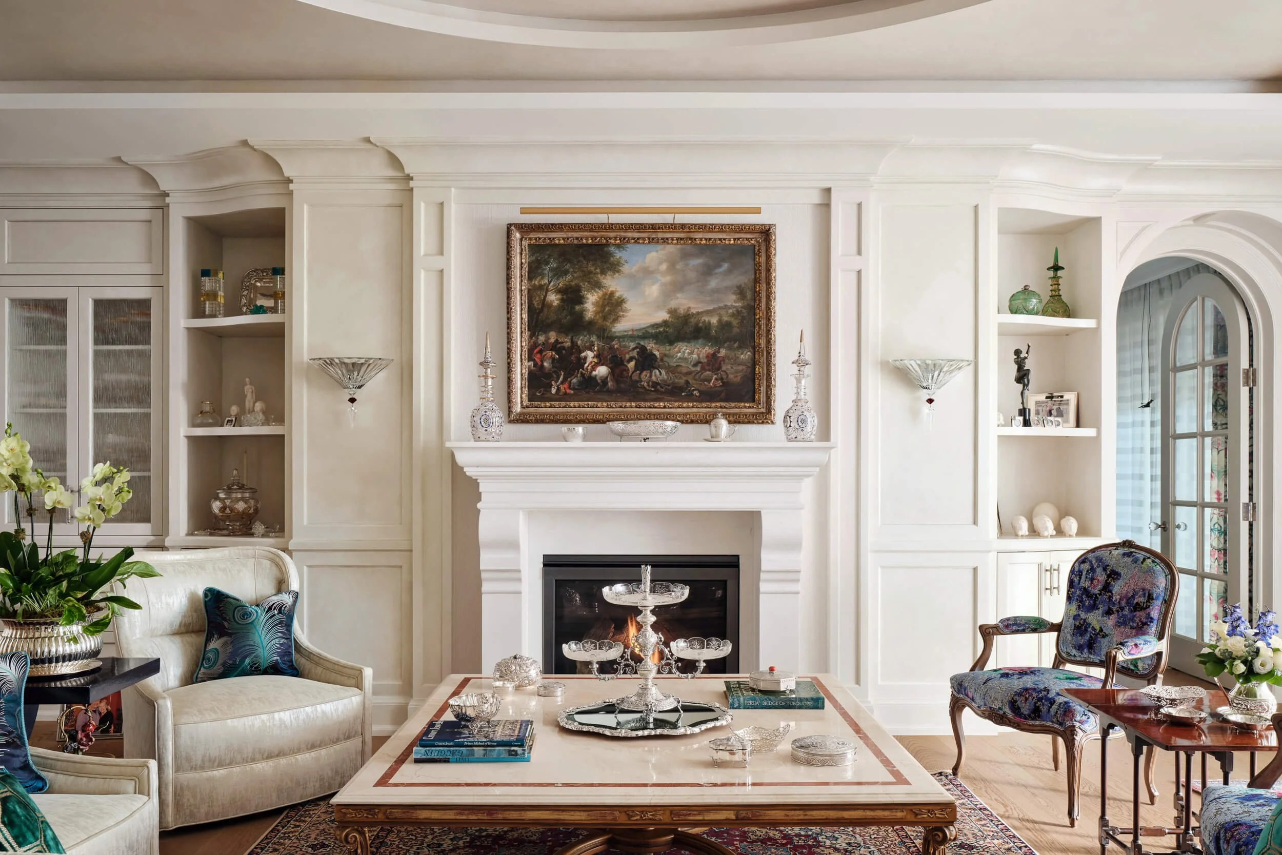Living room with white built-in shelves, a white fireplace with a painting above, and various decorative items. There are upholstered chairs, a coffee table with books and glassware, and plants.