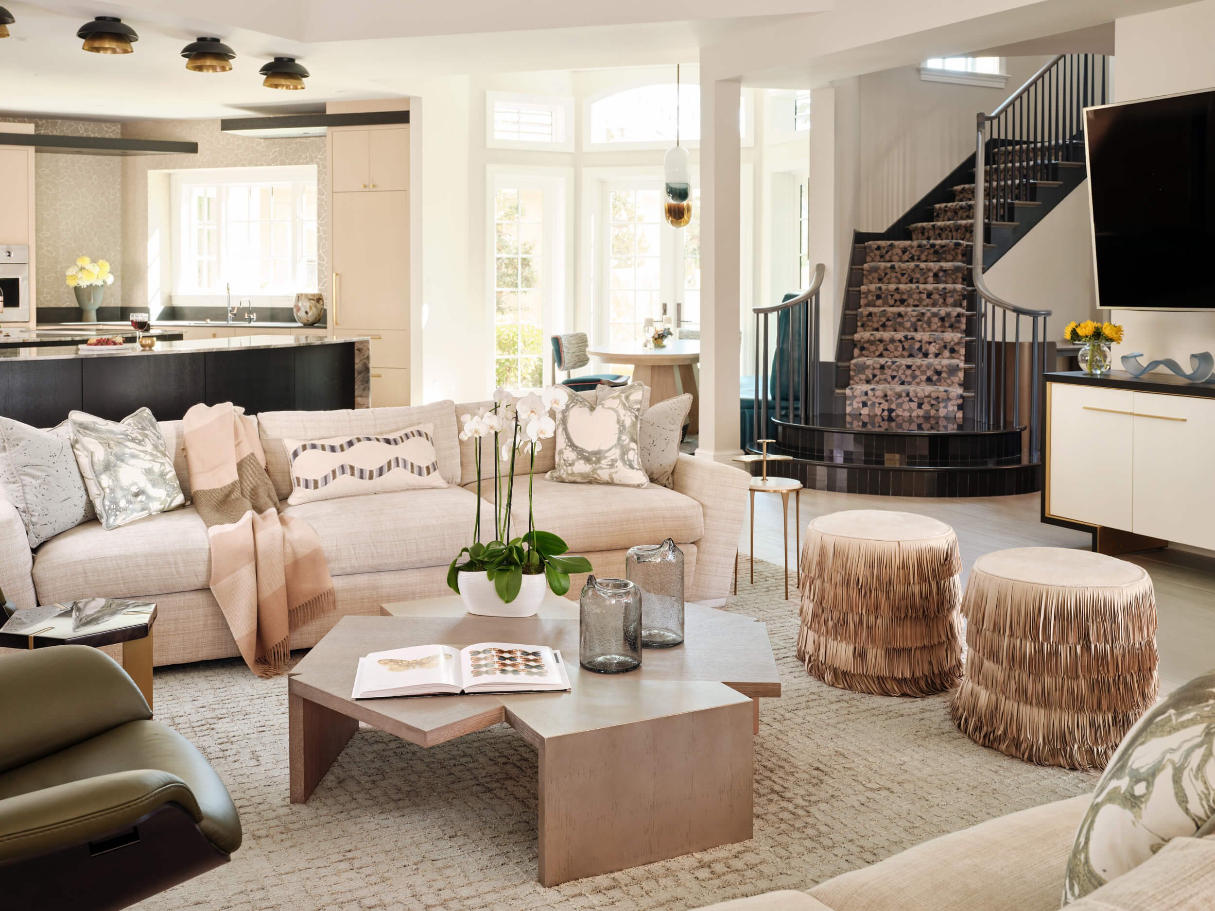 A modern, open-plan living room and kitchen area with beige, white, and brown color schemes. There is a beige sofa with patterned pillows, a wooden coffee table with open book and glass vases, and two fringed stools. The kitchen features black and white cabinetry, a marble island with wine and snacks, and contemporary lighting fixtures. Sunlight streams in through large windows, illuminating the space.