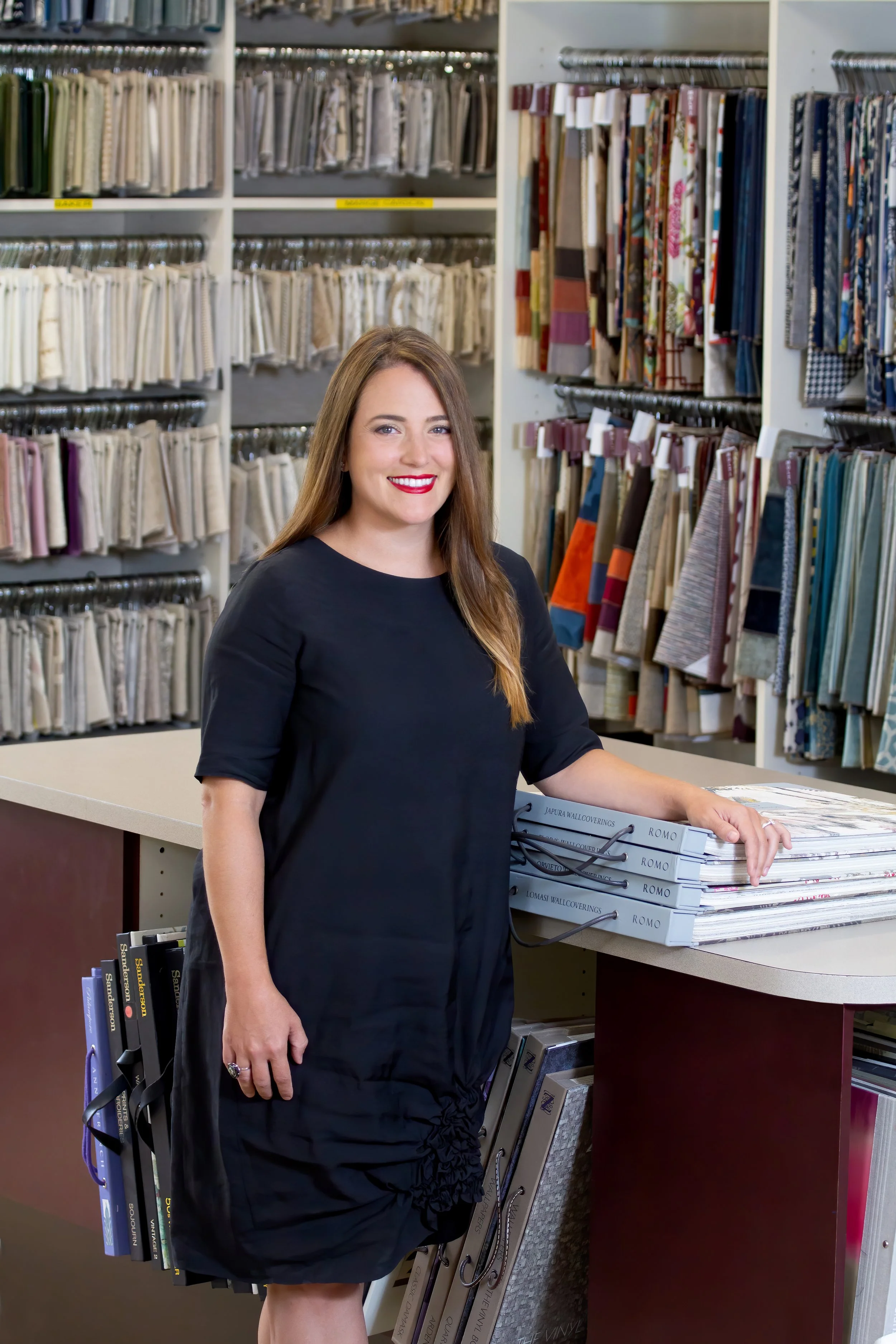 A woman in a black dress with long brown hair and red lipstick standing at a counter in a fabric and wall covering store, with shelves of fabric samples and wallpaper catalogs behind her.