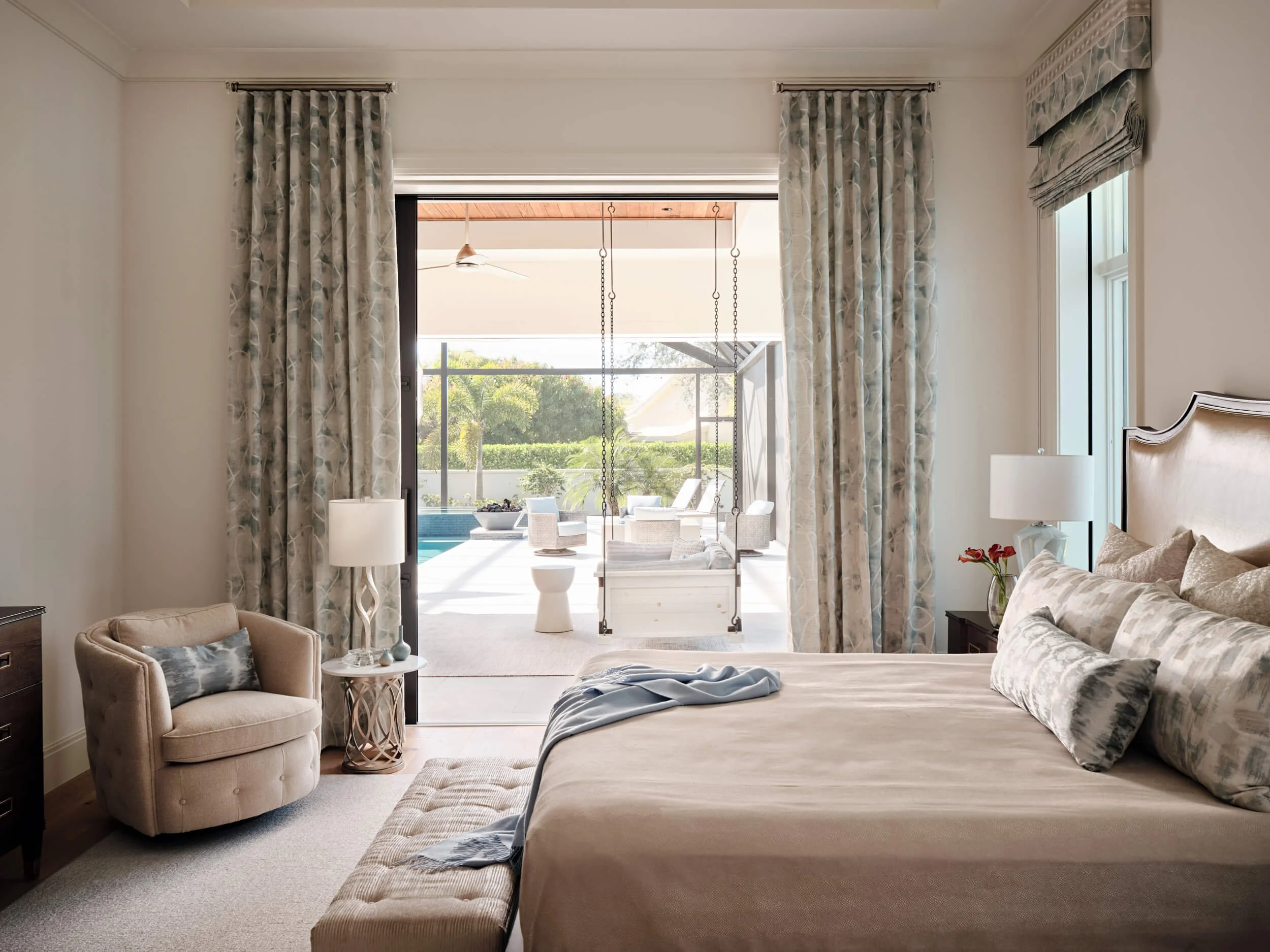 A bedroom with a sliding glass door opening to a patio with outdoor furniture and trees in the background. The room has beige walls, a bed with pillows, a nightstand with a lamp, a plush armchair, and patterned curtains.