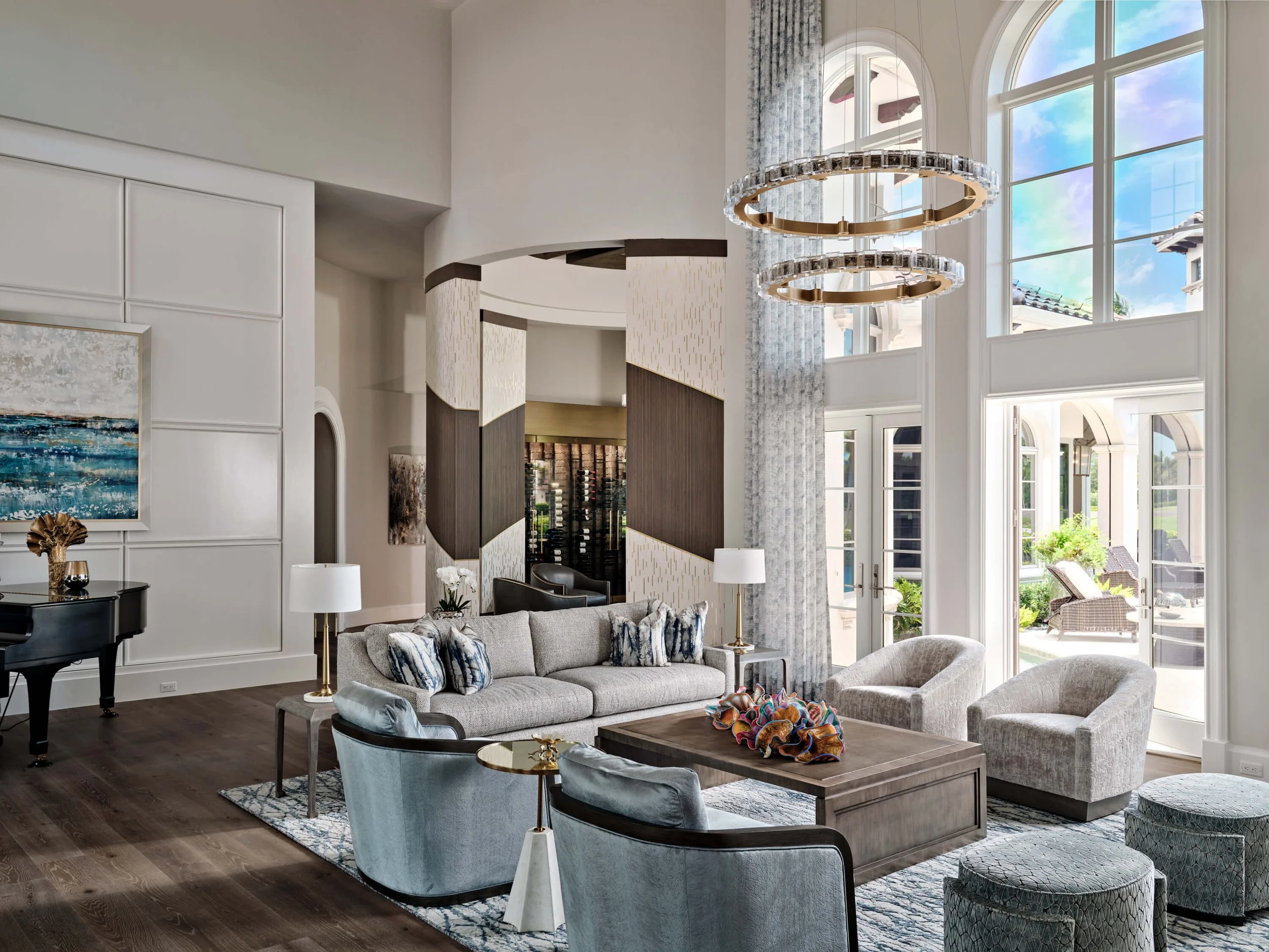 A spacious, modern living room with high ceilings and large arched windows letting in natural light. The room features a light-colored sofa with decorative pillows, a wooden coffee table, and a light-colored armchair. A contemporary, circular chandelier hangs from the ceiling, and there is an abstract painting on the wall. The room has dark wood flooring and is decorated with stylish lamps and accessories.