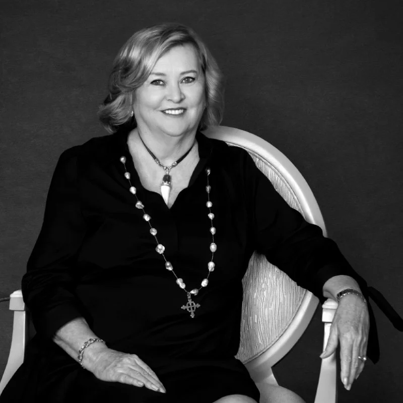 Black and white portrait of a middle-aged woman with short, wavy hair, smiling and sitting in a patterned chair. She is wearing a dark blouse, a long pearl necklace with a crucifix, a bracelet, and a ring. The background is plain and dark.