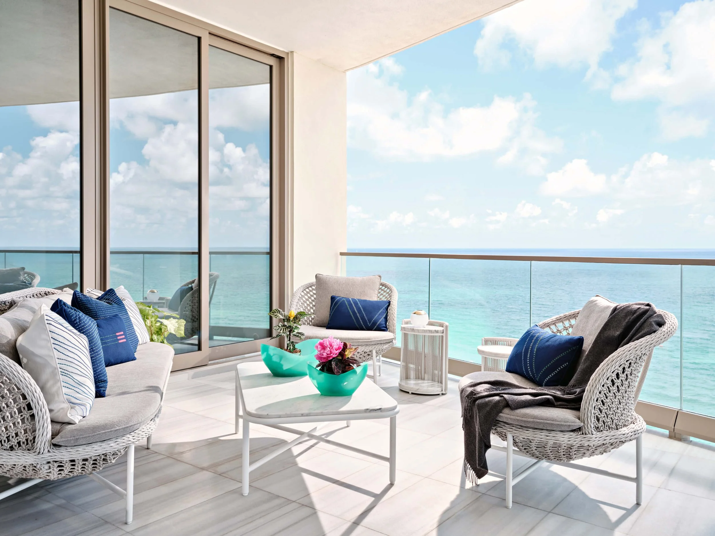 Balcony with white wicker patio furniture including a sofa and chairs with blue and white pillows, a coffee table with a pink flower arrangement, a glass railing, and a view of the blue sky, ocean, and clouds.
