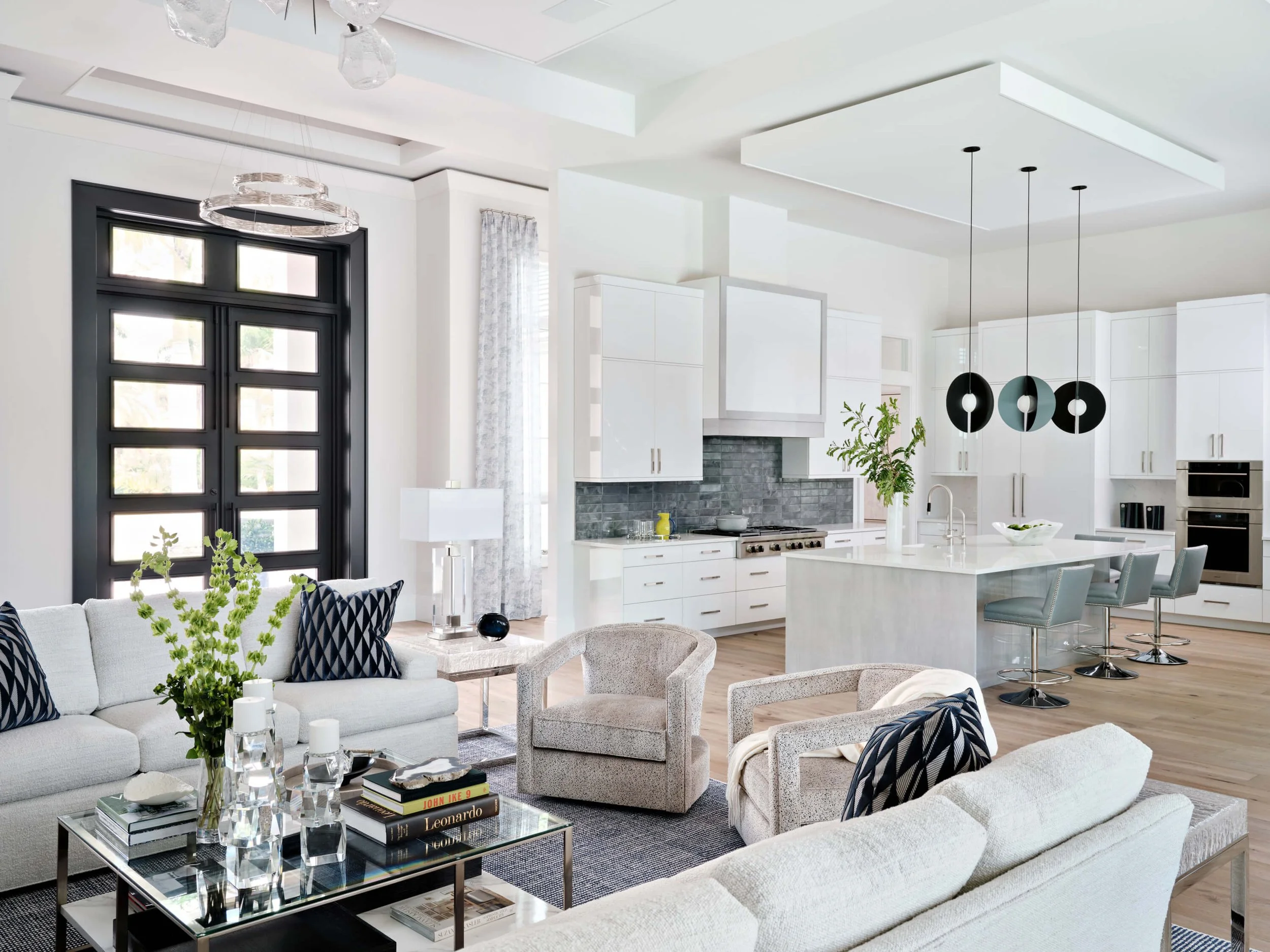 A modern, bright kitchen and living room space with white cabinetry, black-framed front door, hanging black and gray pendant lights, a white sofa with black and white pillows, a glass coffee table with books and a vase of green plants, multiple chair