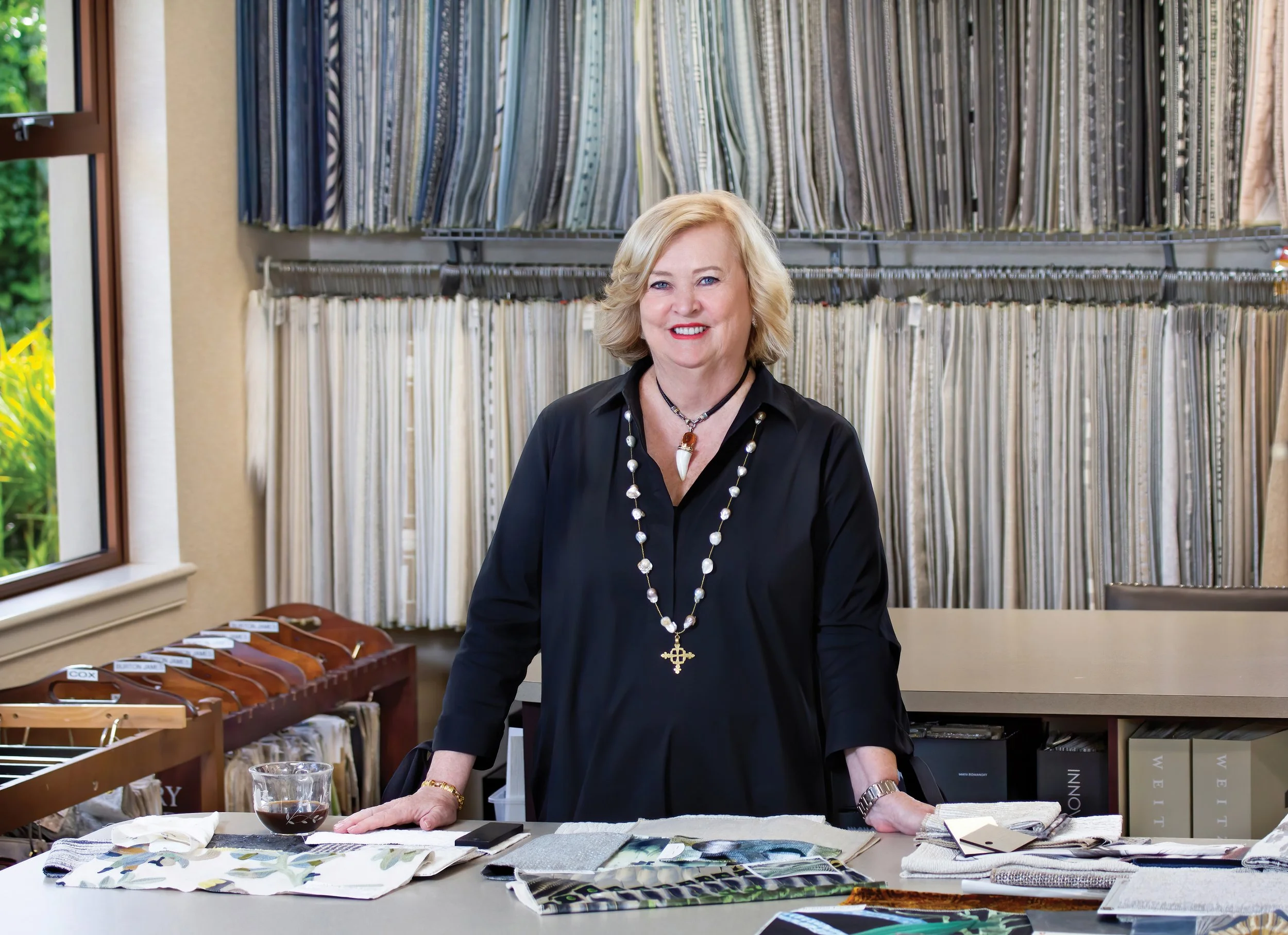 A woman with blonde hair, wearing a black top and pearl necklace, standing behind a table with fabric swatches and a glass of red wine, in a room with fabric samples and a large window.