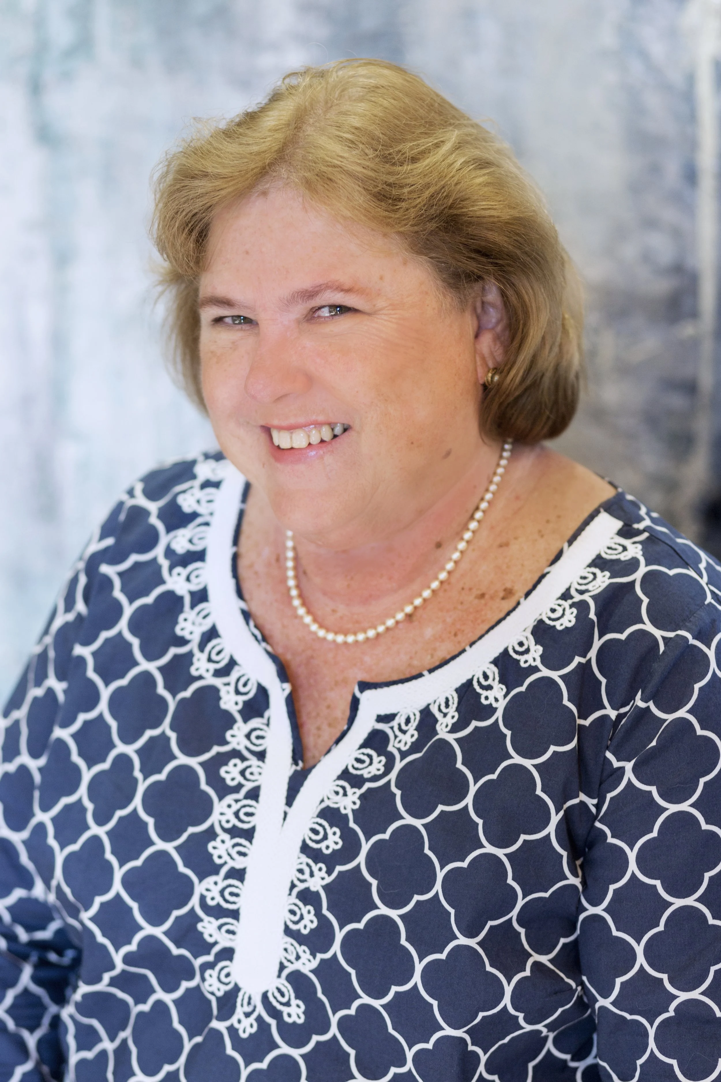 Close-up of a middle-aged woman with short blonde hair, wearing a blue and white patterned dress, pearl necklace, and earrings, smiling with her face slightly turned to the side.