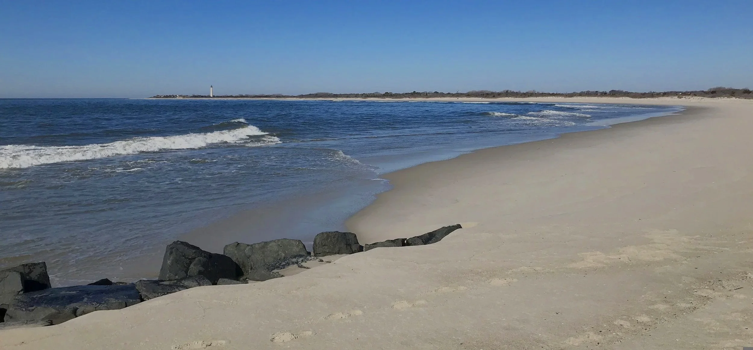 A sandy beach with rocks in the foreground, gentle waves in the ocean, and a lighthouse in the distance under a clear blue sky.