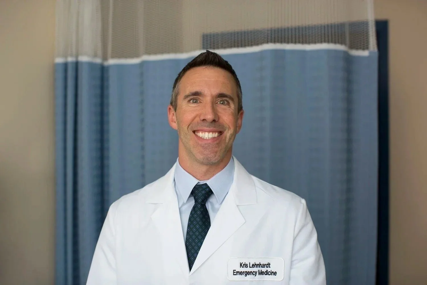 Smiling male doctor in white coat with a name tag reading 'Kris Lehnhardt Emergency Medicine' poses for a photo in a hospital setting.