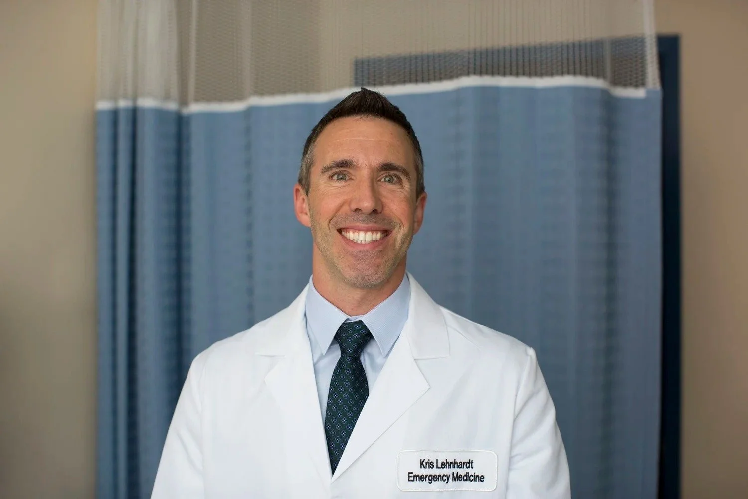 Smiling male doctor in white coat with a name tag reading 'Kris Lehnhardt Emergency Medicine' poses for a photo in a hospital setting.