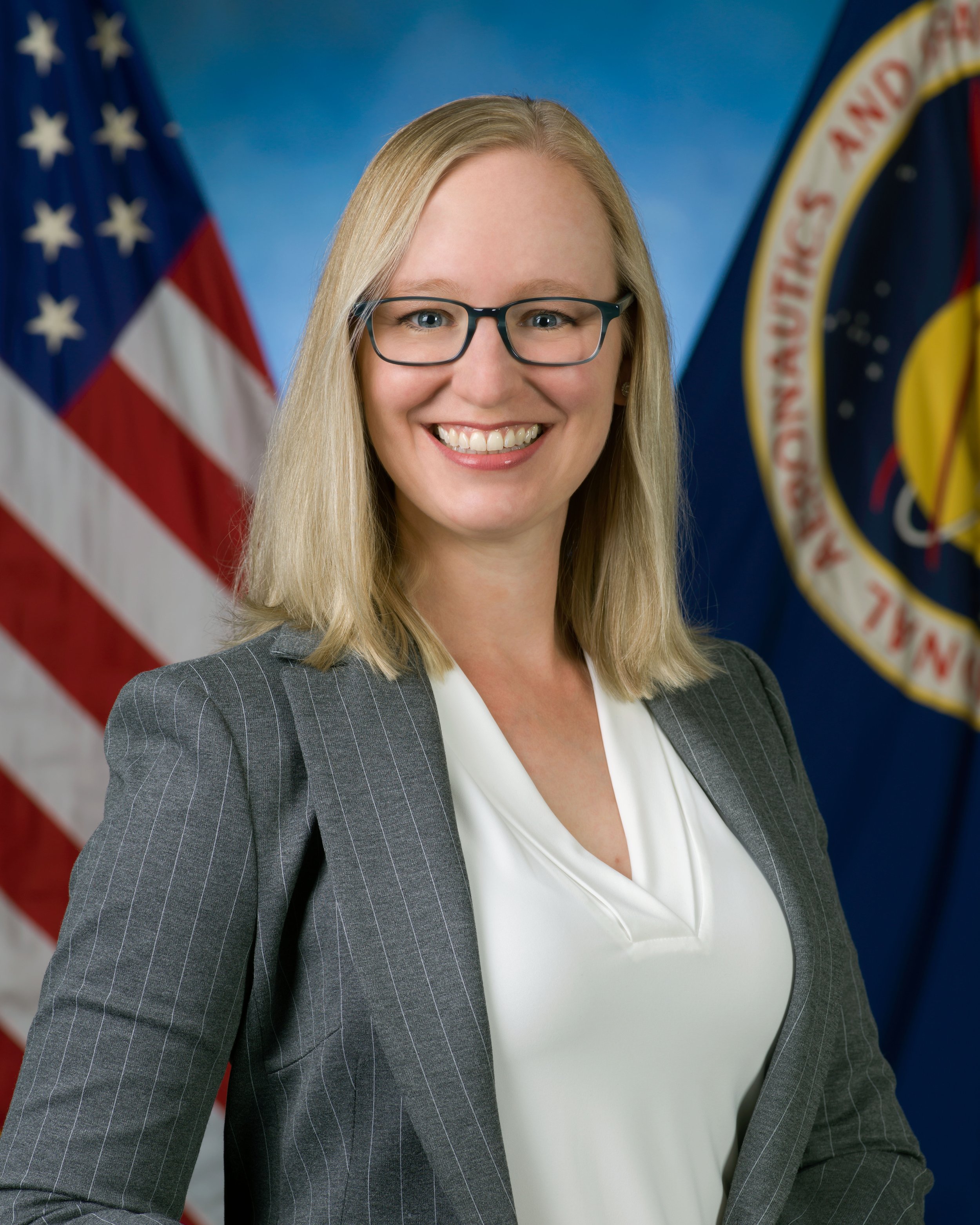 A woman with blonde hair and glasses wearing a gray business suit stands in front of American and NASA flags.