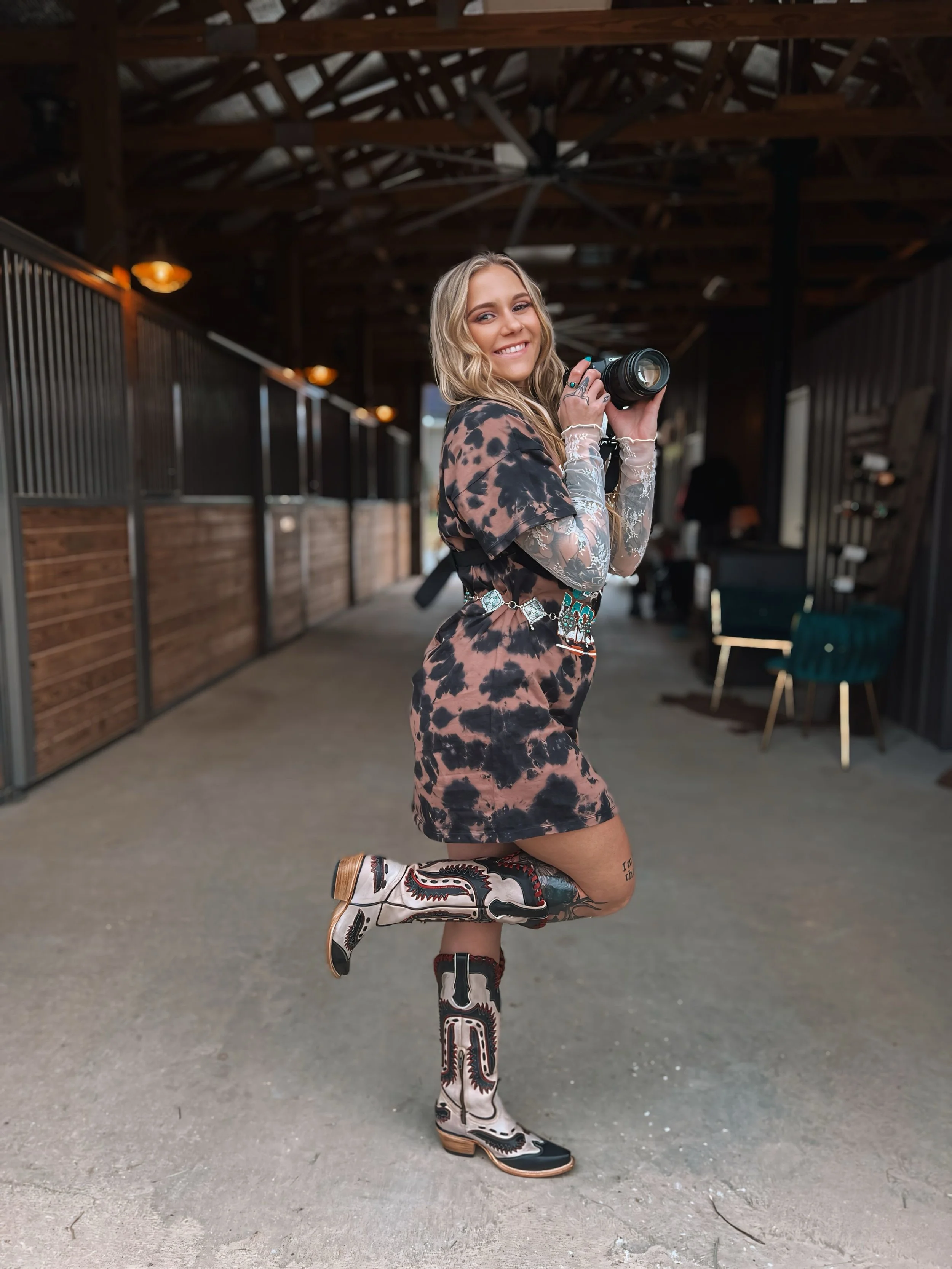 Young woman with long blonde wavy hair holding a camera, smiling, standing on one leg with the other foot bent backward, wearing a tie-dye dress, lace sleeve tattoos, and cowboy boots, in a rustic, covered barn or stable.