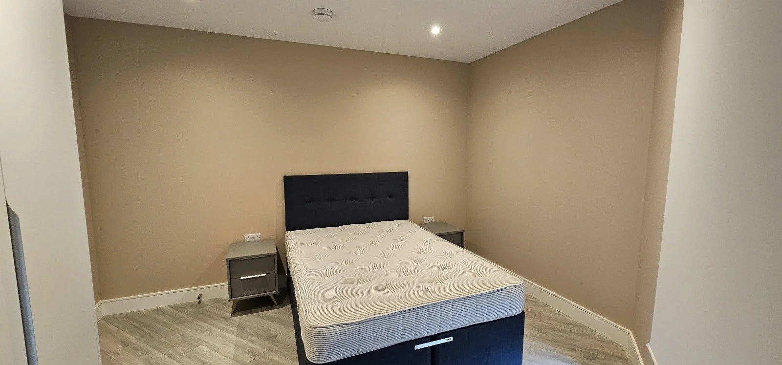 A bedroom with beige walls, a bed with a black headboard and a bare mattress, two gray nightstands on either side of the bed, and a ceiling light.