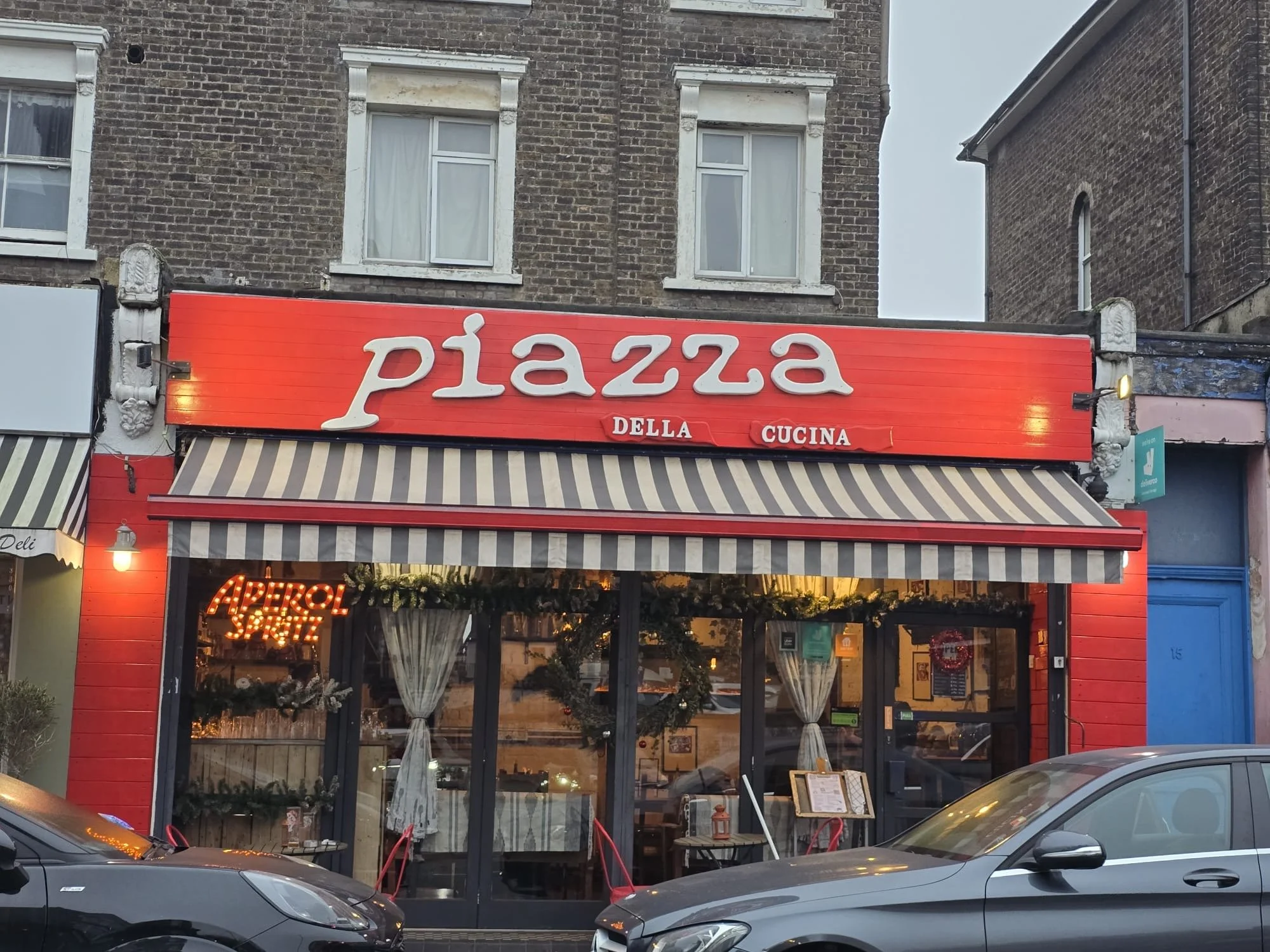 The exterior of a restaurant called 'Piazza Della Cucina' with large windows, white curtains, and red and white striped awning. The restaurant is situated in a brick building.