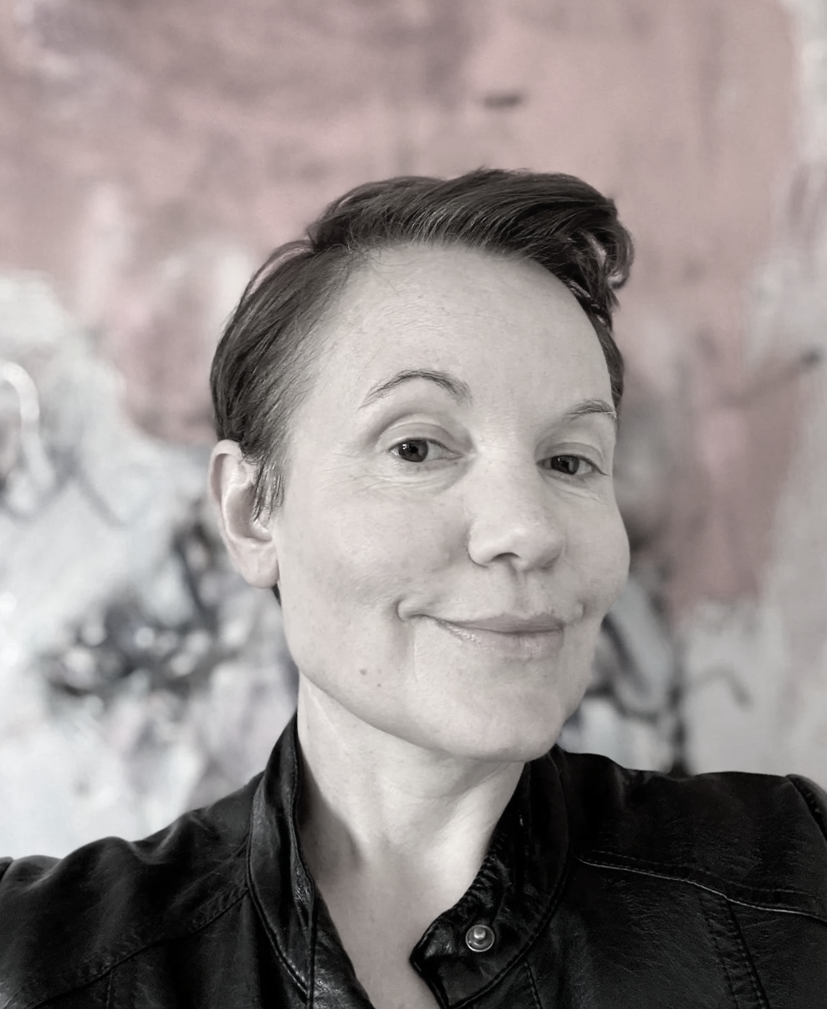 A woman taking a selfie with a subtle smile, short hair, wearing a leather jacket, against a blurred background.