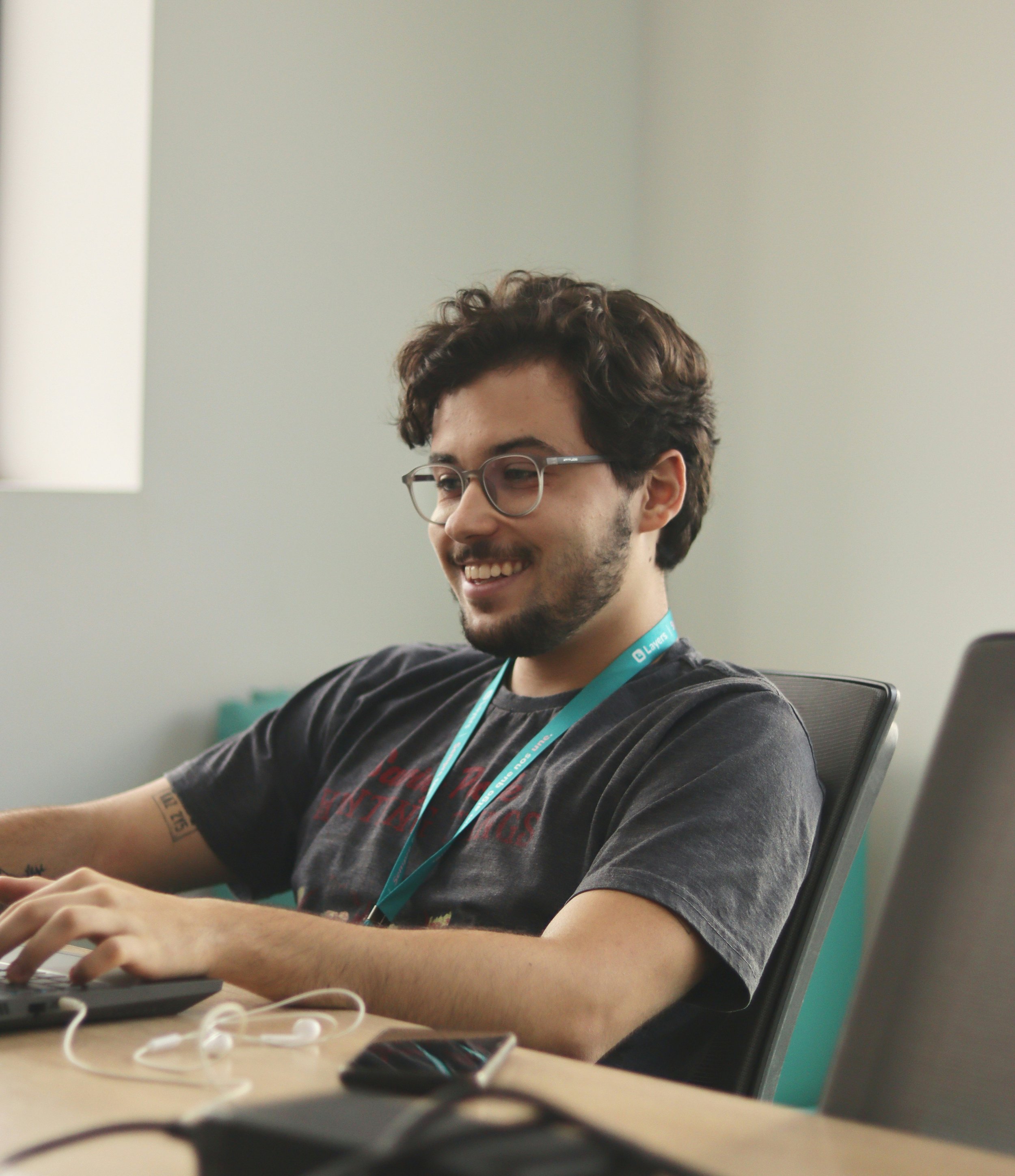Joven hombre con gafas y barba, sonriendo mientras trabaja en una computadora en una oficina moderna.