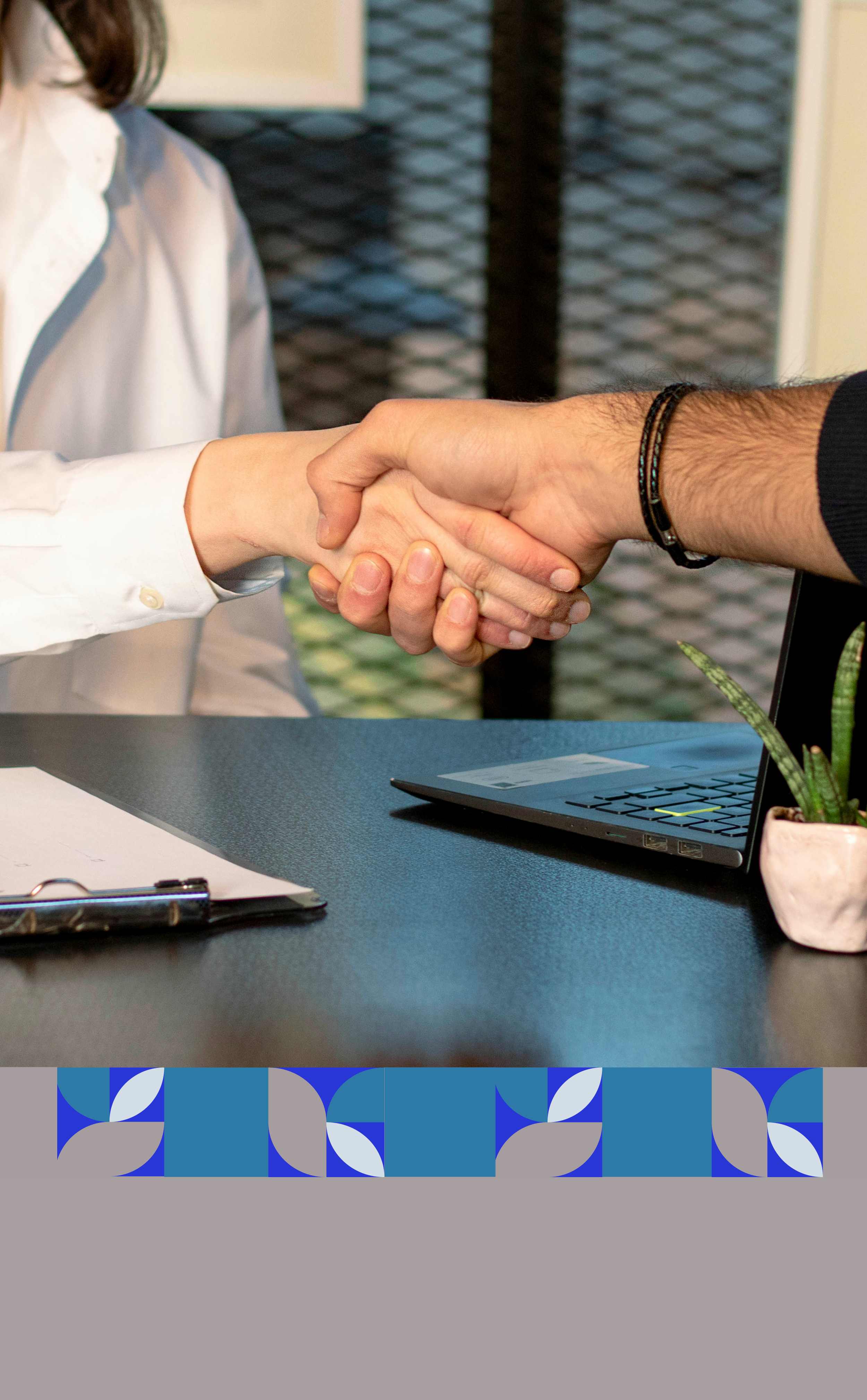 Persona y otra persona estrechándose la mano en una reunión, en una mesa con una computadora portátil, un portapapeles y una planta.