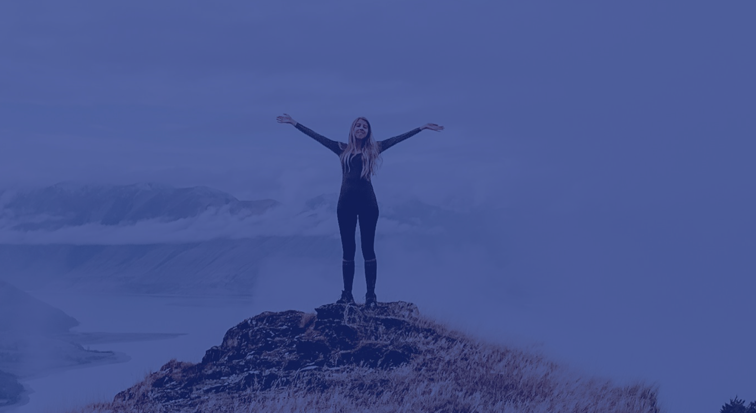 Mujer de pie sobre una roca con los brazos abiertos en un paisaje de montaña con nubes y agua en el fondo.