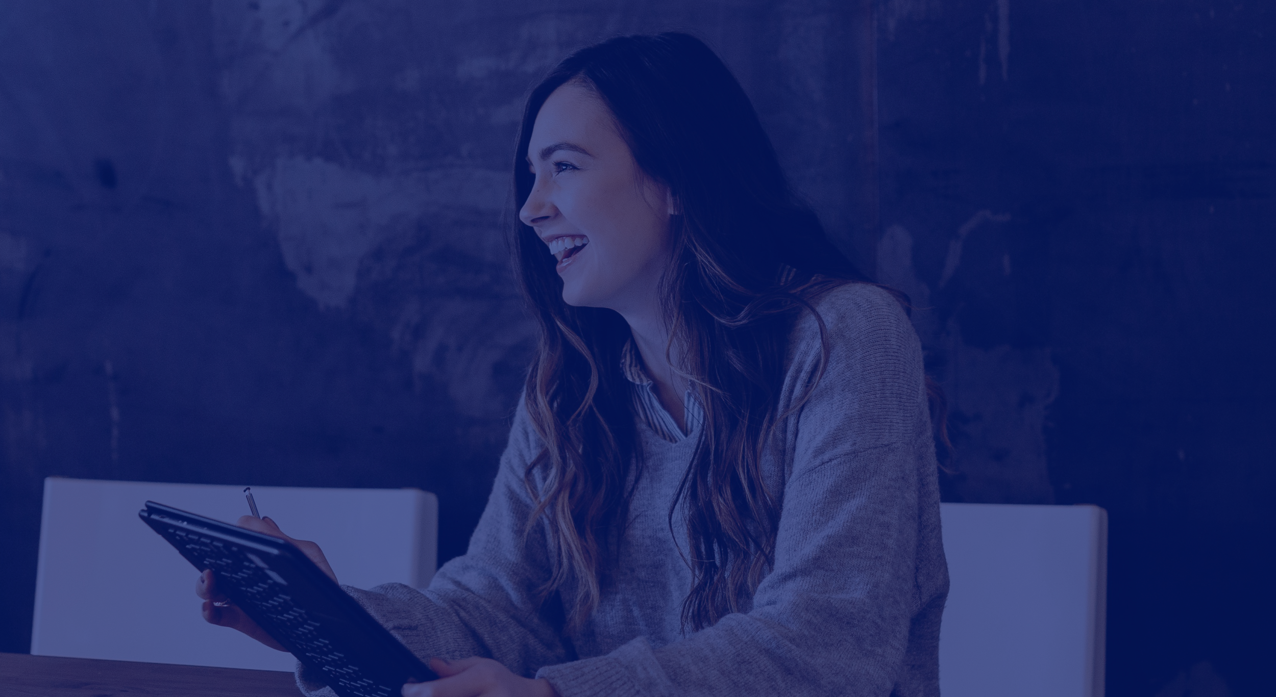 Una mujer riendo y sonriendo, sentada en una mesa, sosteniendo un teléfono inteligente, con un fondo de pared oscura.