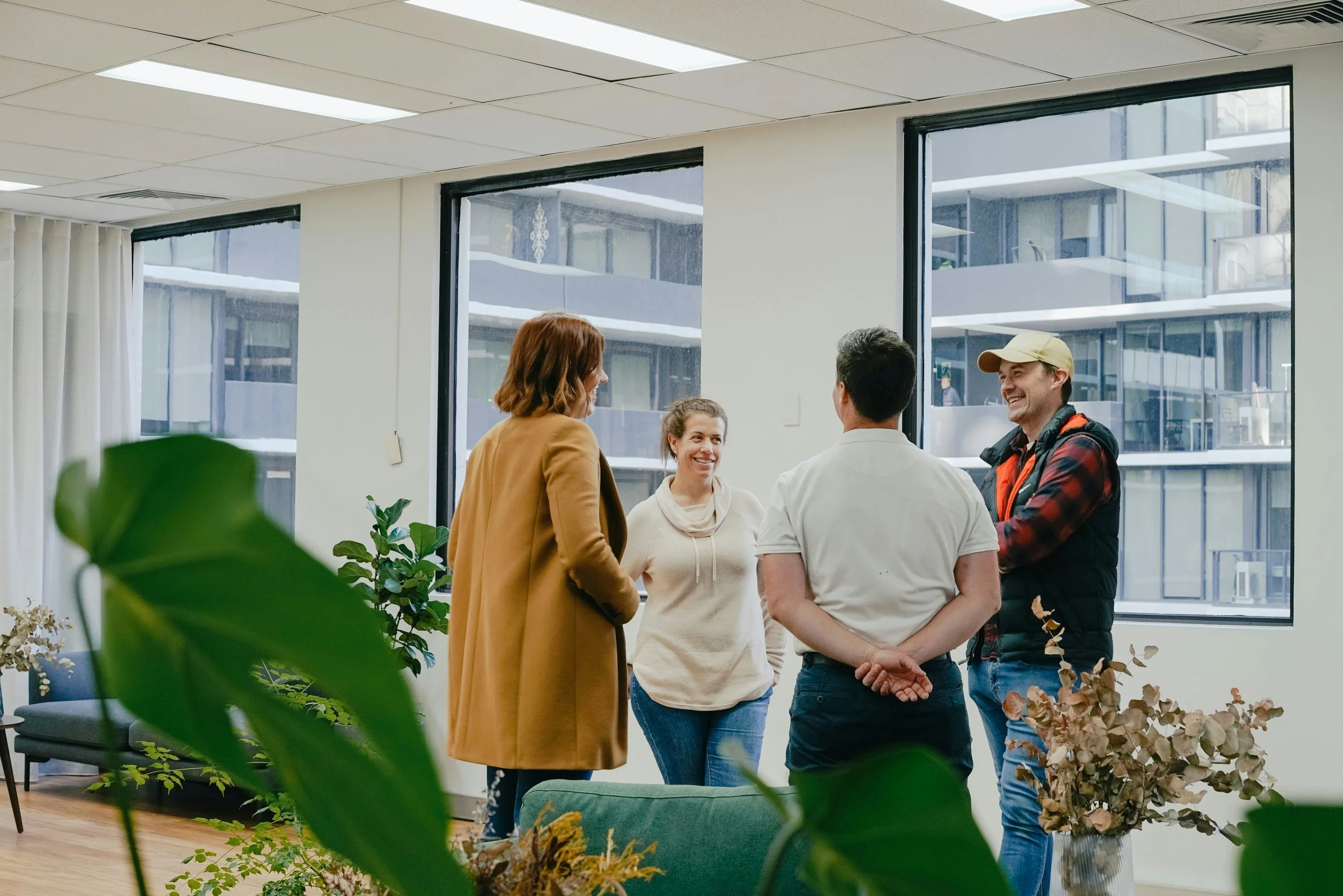 Grupo de cuatro personas conversando en una oficina con ventanas grandes y plantas decorativas.