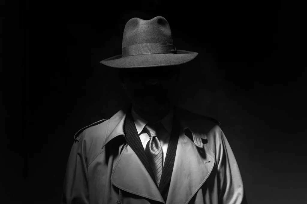 A person wearing a wide-brimmed hat, trench coat, dress shirt, and tie, standing in a dark environment with only faint lighting highlighting the outline.