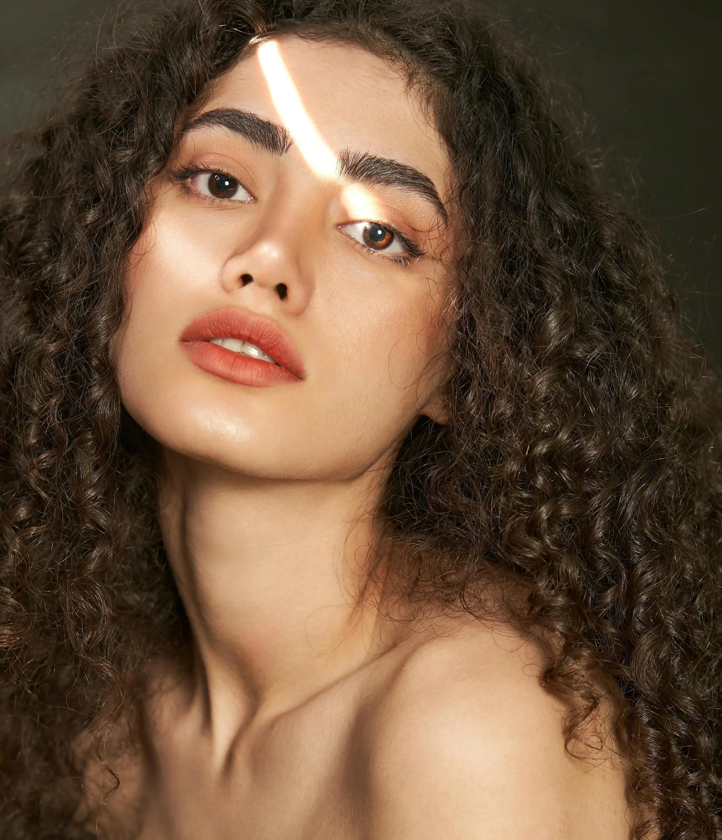 A close-up of a young woman with curly brown hair and light makeup, looking at the camera.