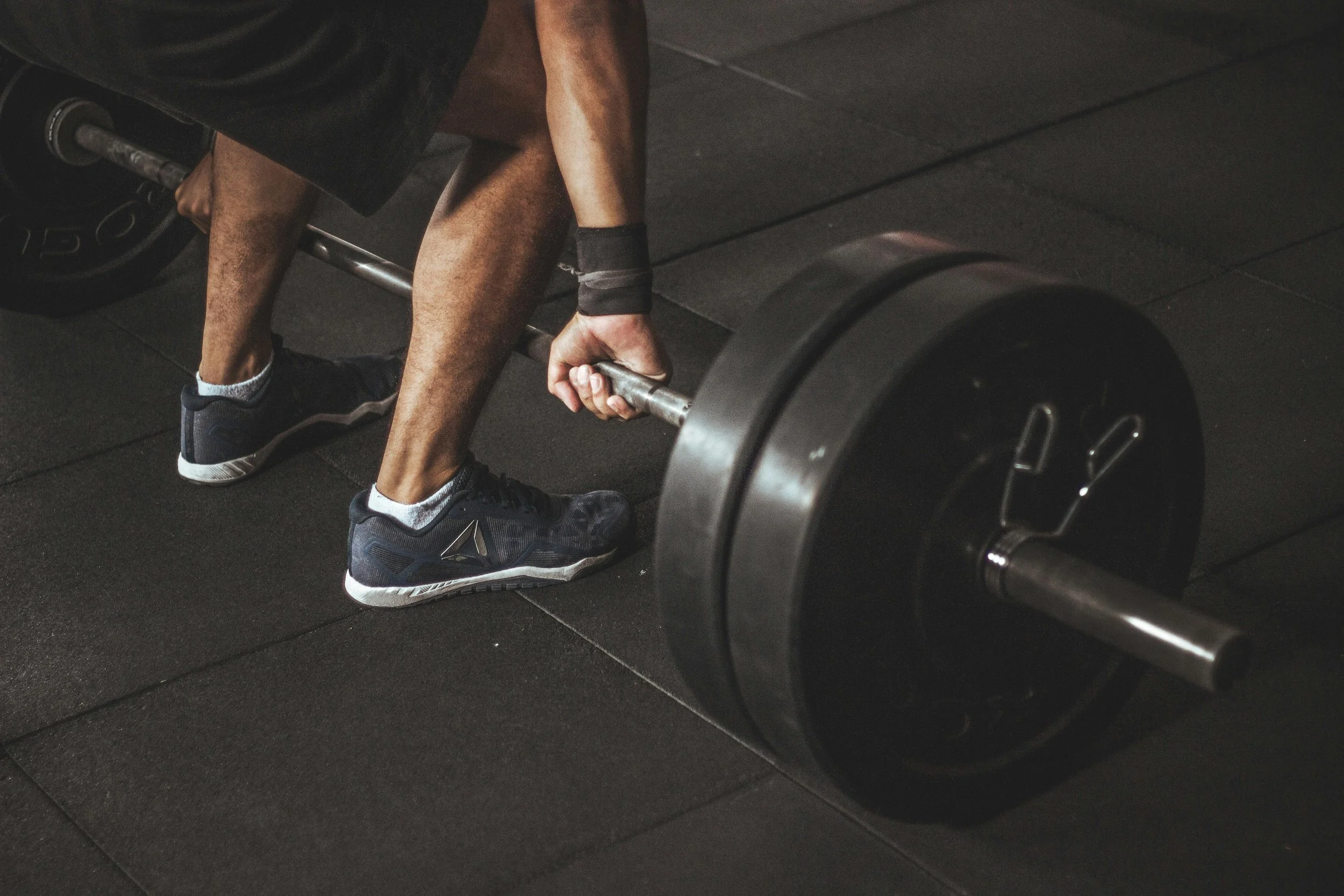 A person in black athletic shoes and shorts bending down and gripping a barbell on the floor in a gym. The barbell has large weight plates on each end.