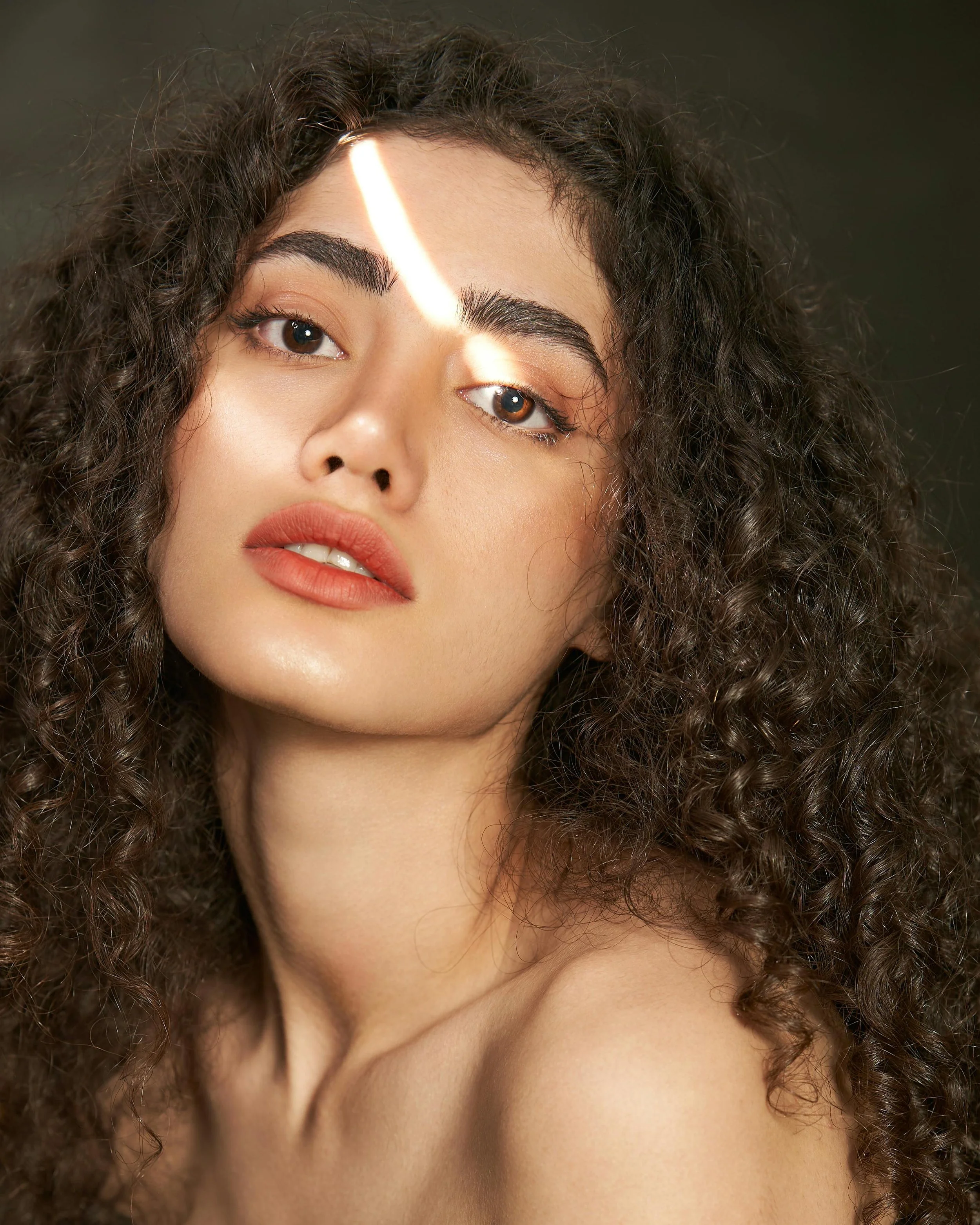 A close-up of a young woman with curly brown hair and light makeup, looking at the camera.