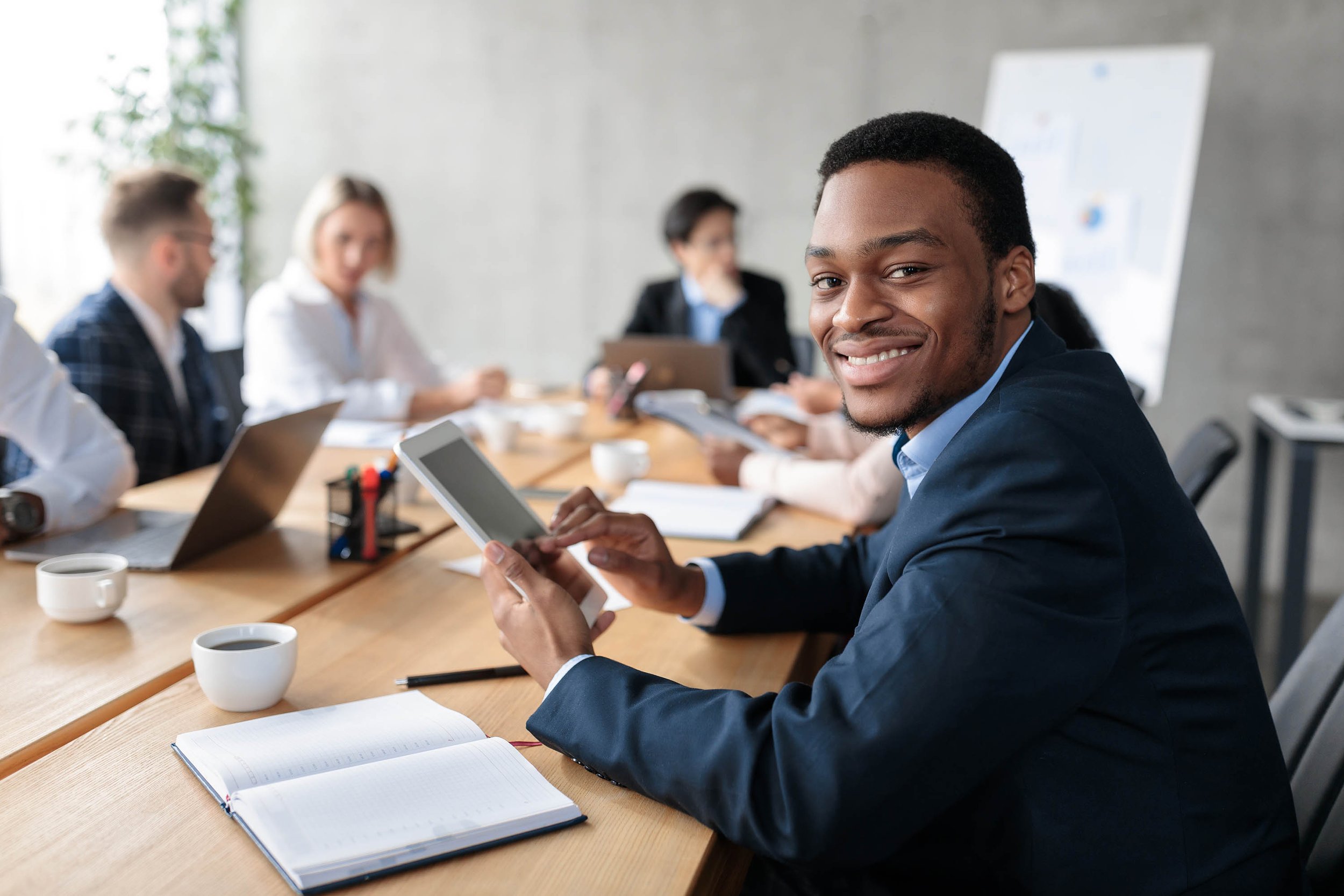 Man sitting at a conference table with other individuals blurred in the background.