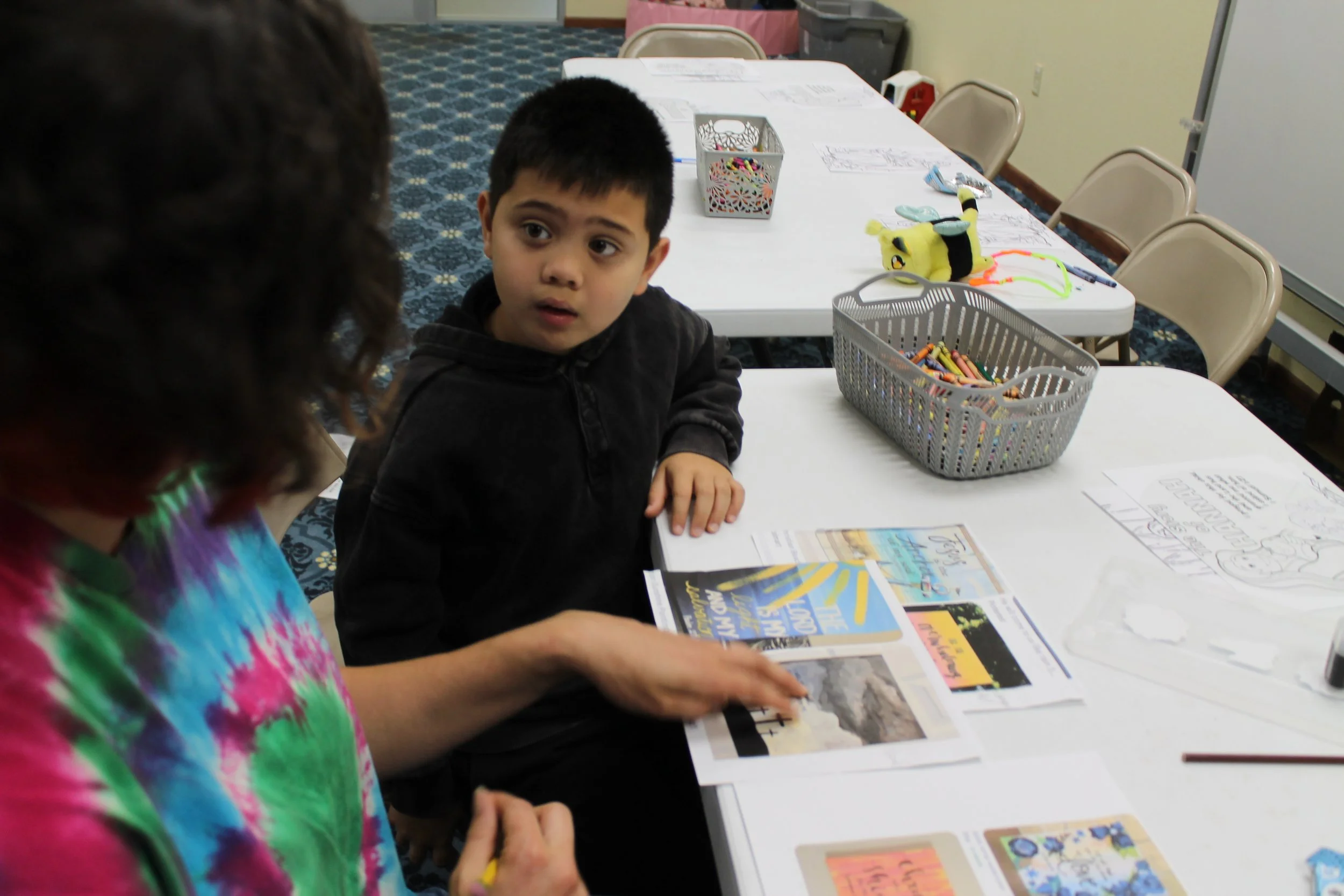 A woman and a young boy sitting at a table, looking at a bible lesson activity, in a room with multiple chairs and arts and crafts supplies on the table.