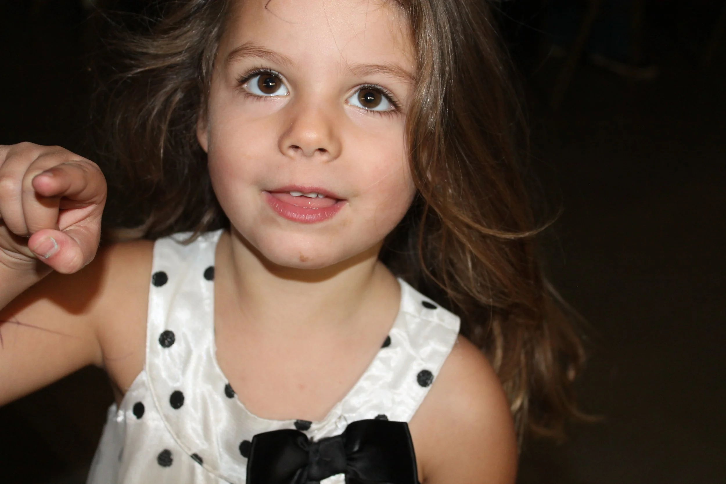 Close-up of a young girl with brown hair, brown eyes, and light skin, wearing a white dress with black polka dots and a black bow, looking up and slightly smiling.