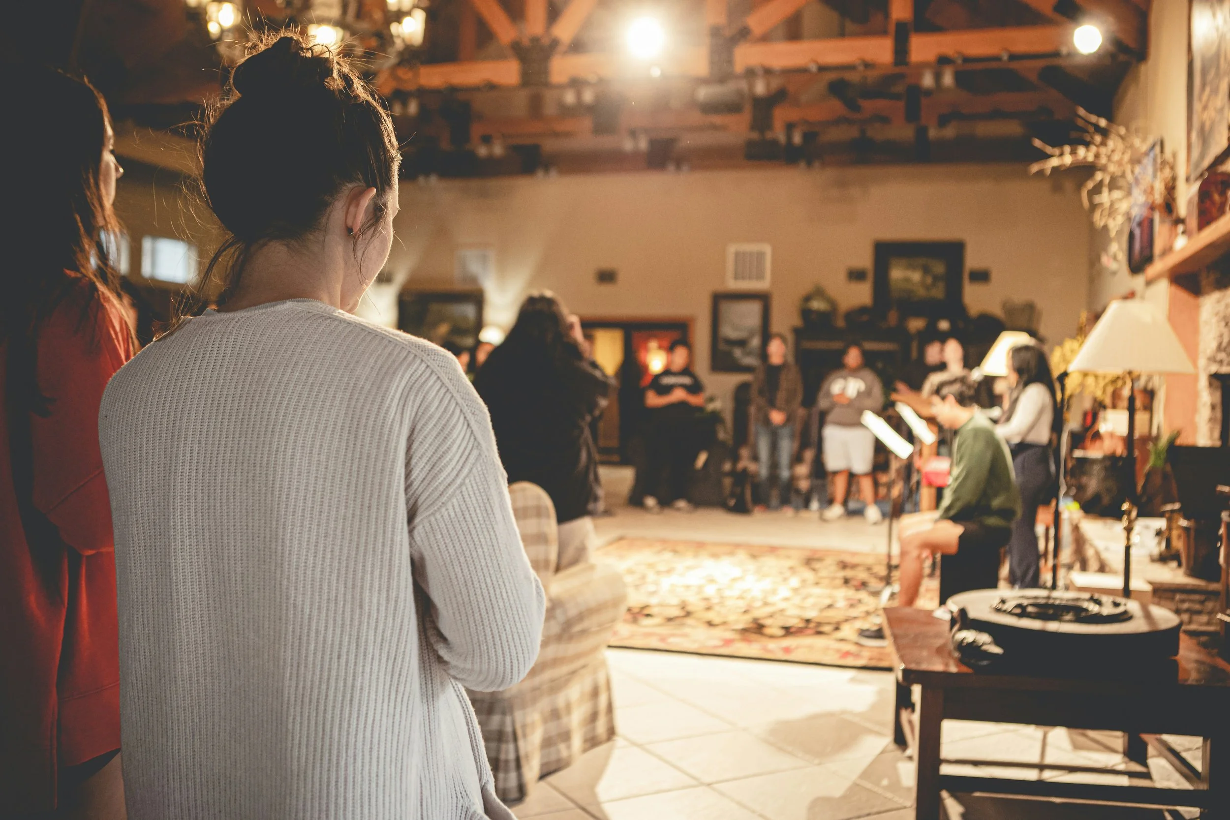 Group of people singing and playing music in a cozy living room with warm lighting, framed pictures, and a patterned rug.
