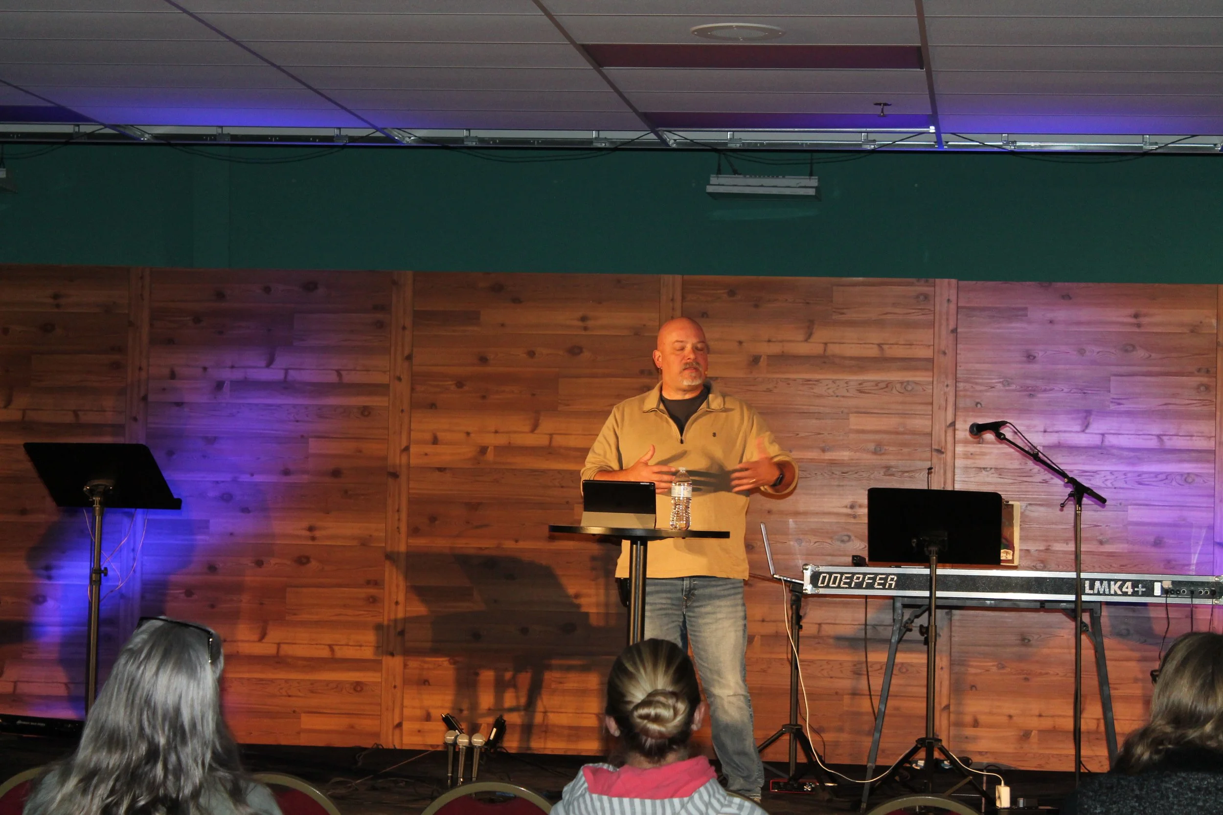Russ McElveen preaching on stage with wooden backdrop, surrounded by musical equipment and audience members.