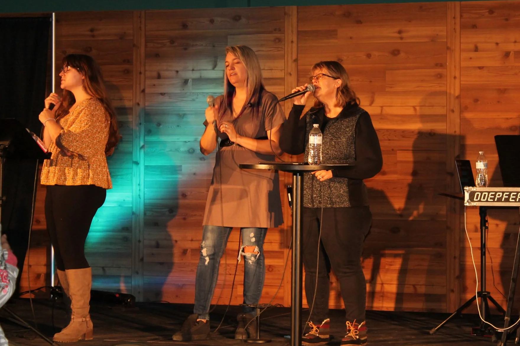 Three women on stage, each holding a microphone. They are standing in front of a wooden-paneled wall, with stage lighting casting shadows. The woman on the right is standing behind a small round table with a water bottle, and there is a keyboard on a stand to the far right.