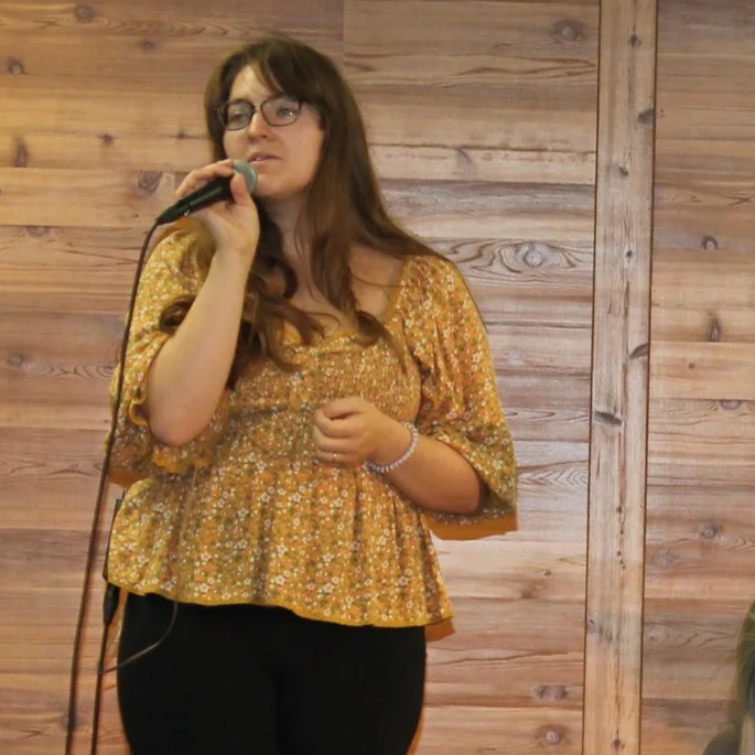 A woman with long brown hair and glasses is holding a microphone and singing in front of a wooden wall.