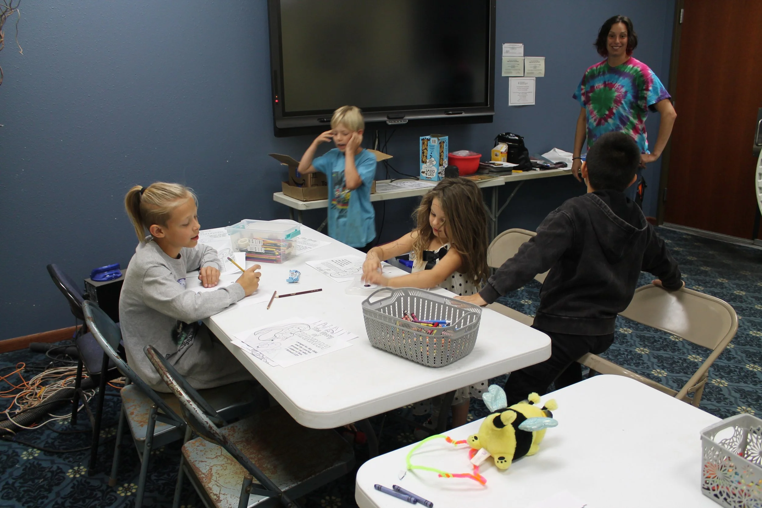 Children engaged in a craft activity at a table with an adult supervising in a room with blue walls and a large digital display monitor.