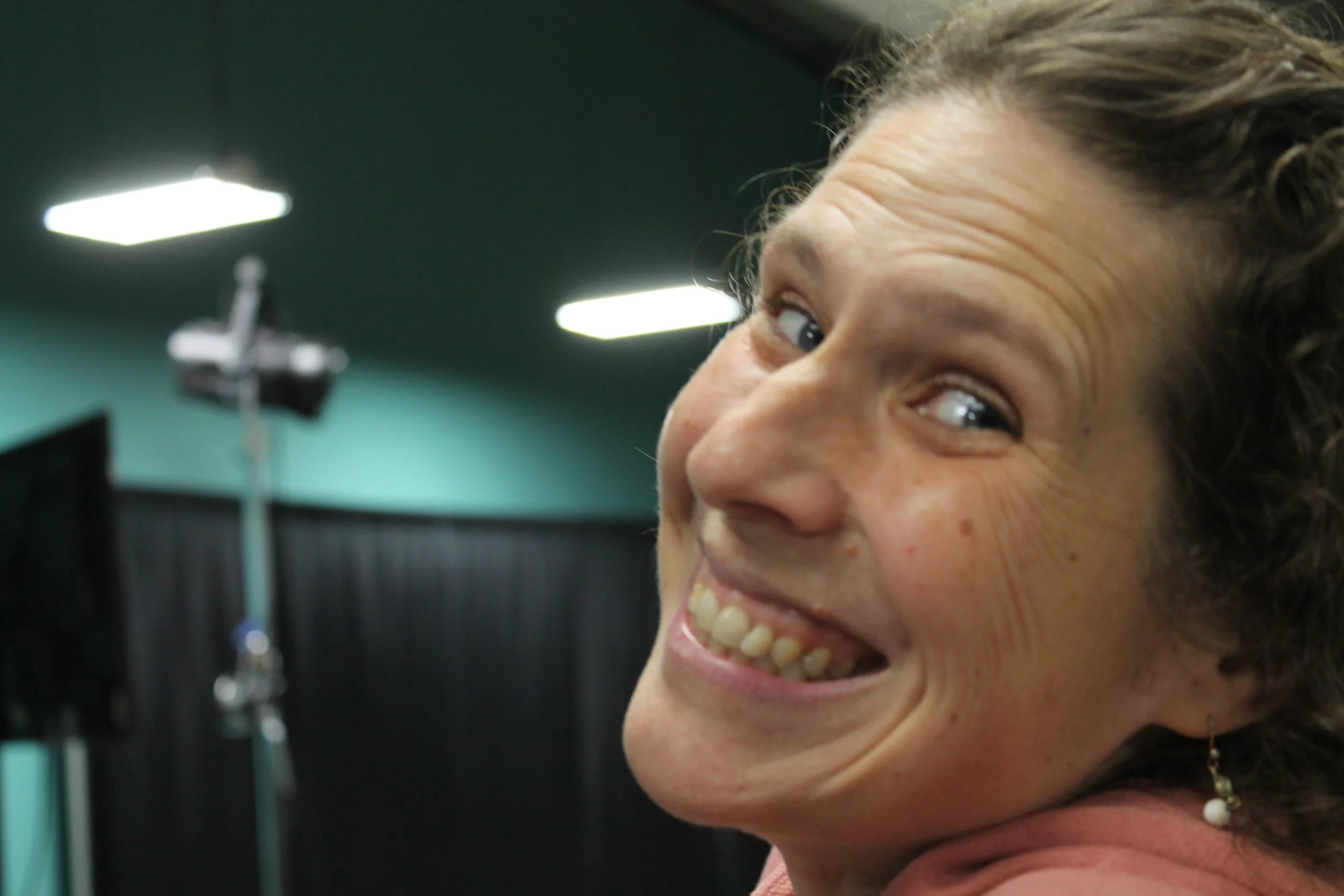Close-up of a woman smiling with visible teeth, in an indoor setting with dark curtains, equipment, and bright lights in the background.