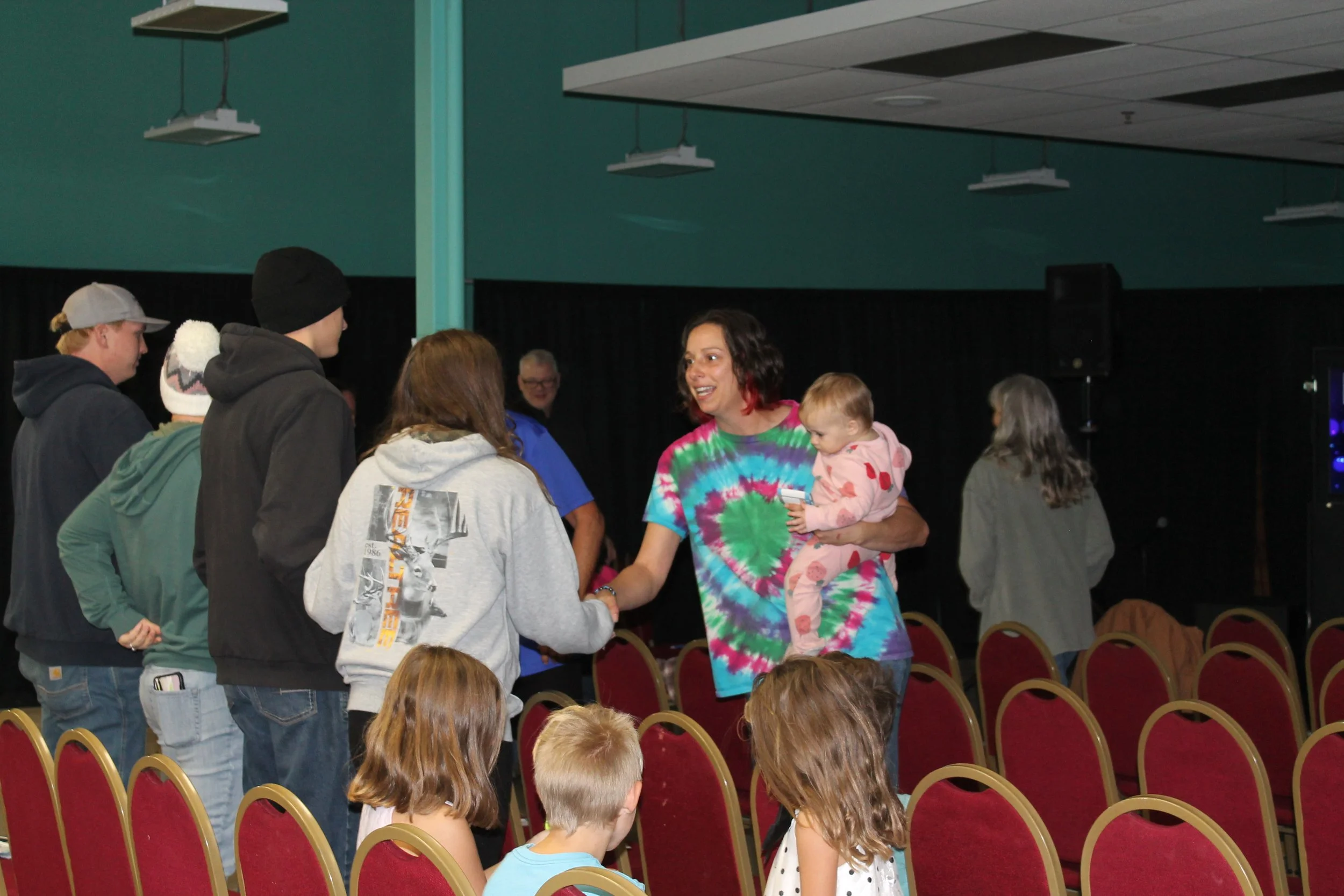 A woman with short dark hair wearing a colorful tie-dye shirt is holding a young girl in pajamas and shaking hands with a couple inside a room with black curtains and red chairs.